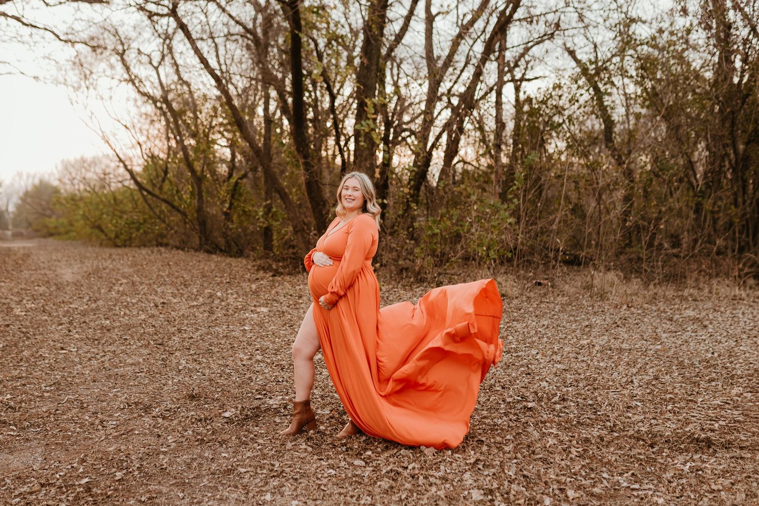 Pregnant woman in flowing orange gown poses outdoors with dress billowing in the wind.