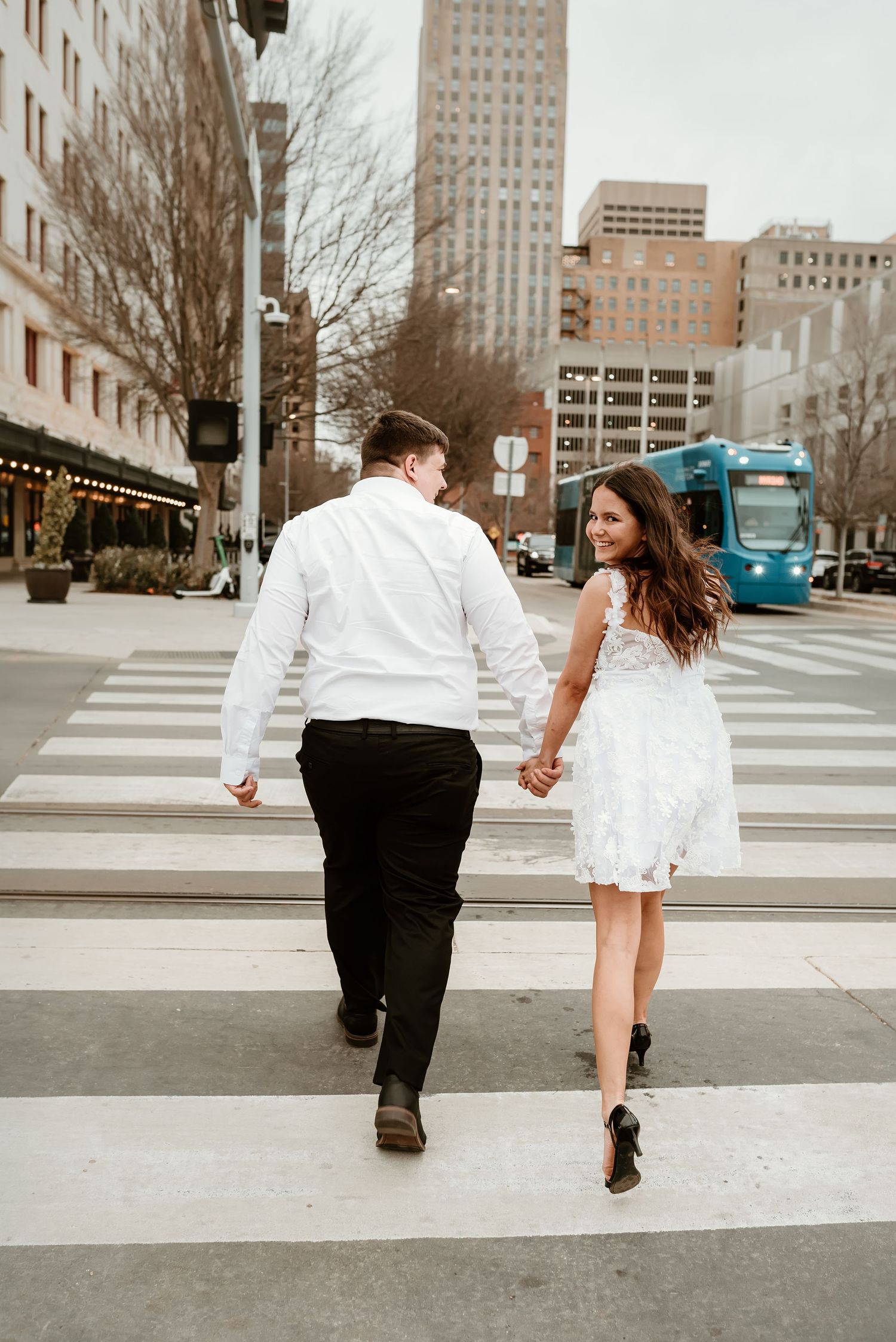 Couple holding hands crossing a city street crosswalk, woman in white lace dress.