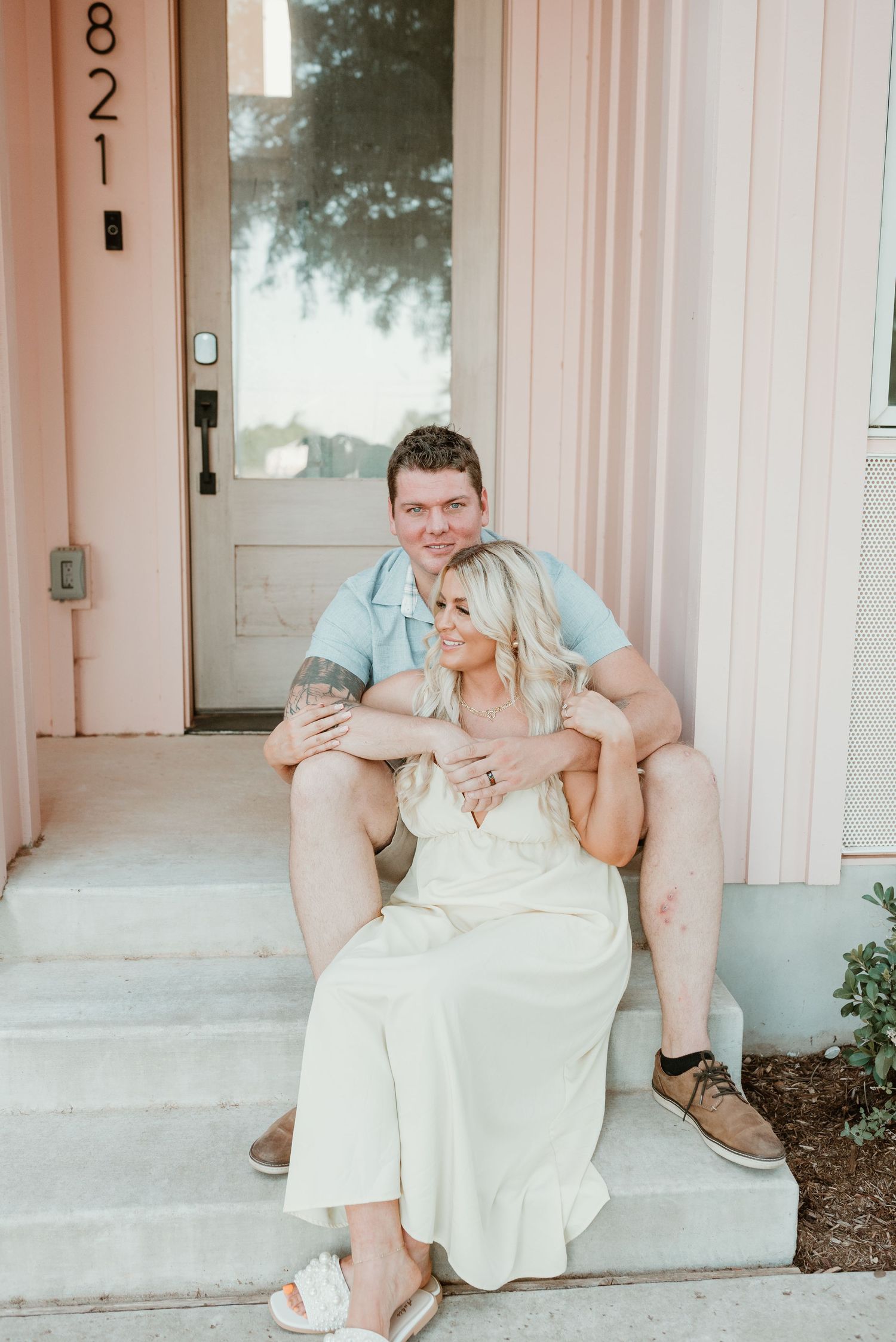 Couple sitting on front porch steps of a pink building, man holding woman from behind warmly.