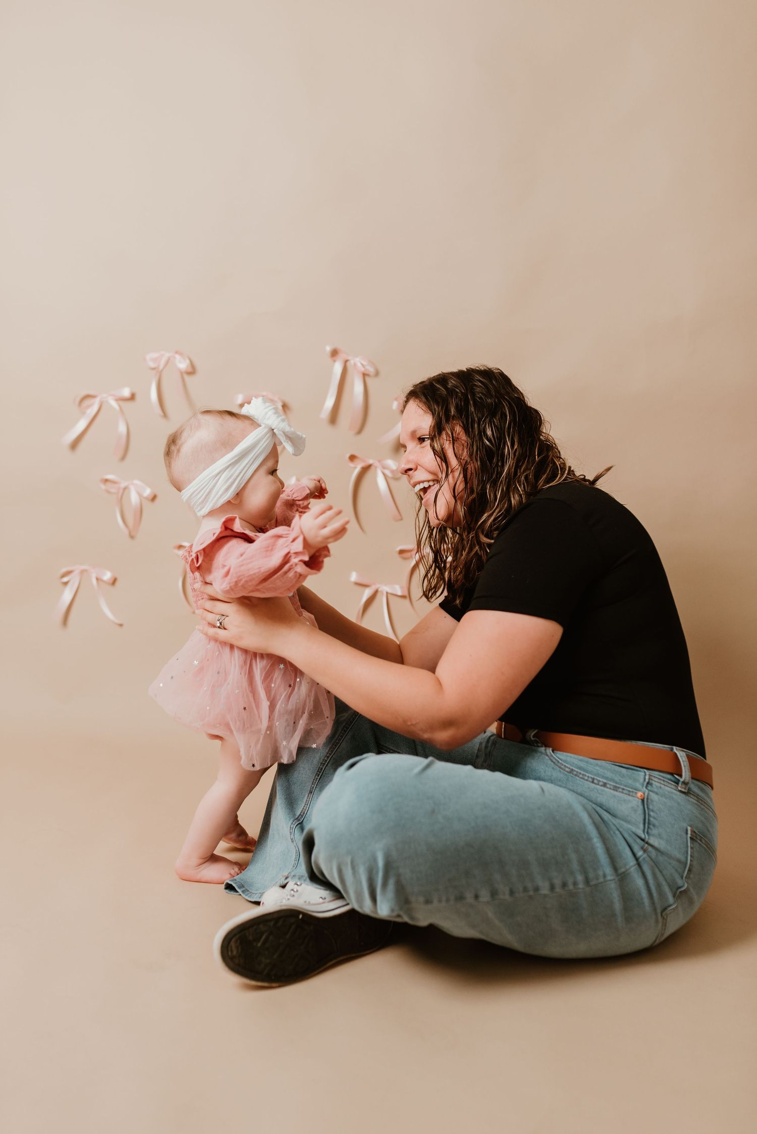 Mother in black top and jeans sits on floor holding baby girl in pink tutu with floating pink bows backdrop.