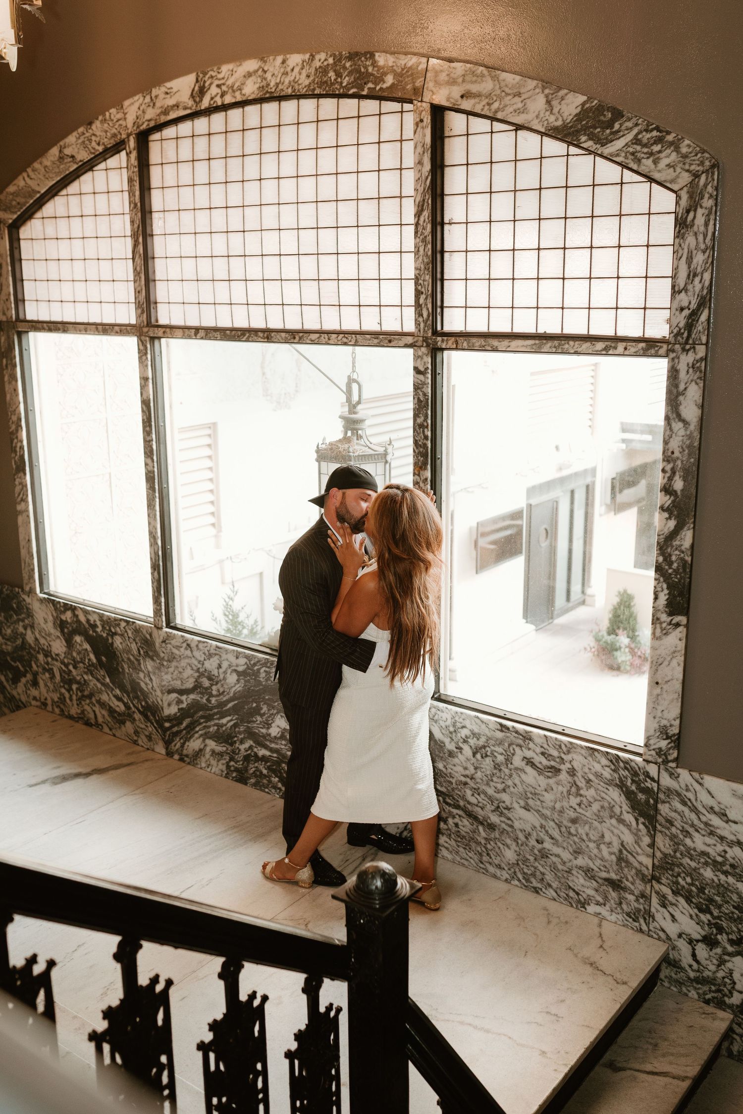 Couple sharing a romantic kiss on a staircase landing by an arched window.