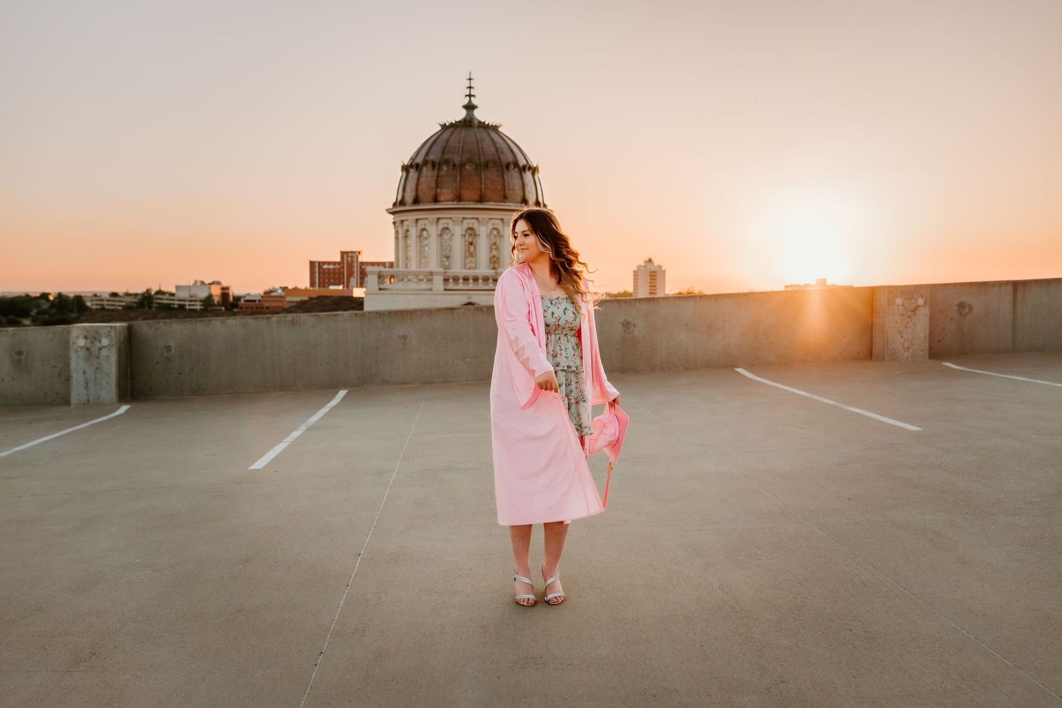 Woman in pink coat standing on rooftop parking lot with domed building and sunset skyline.