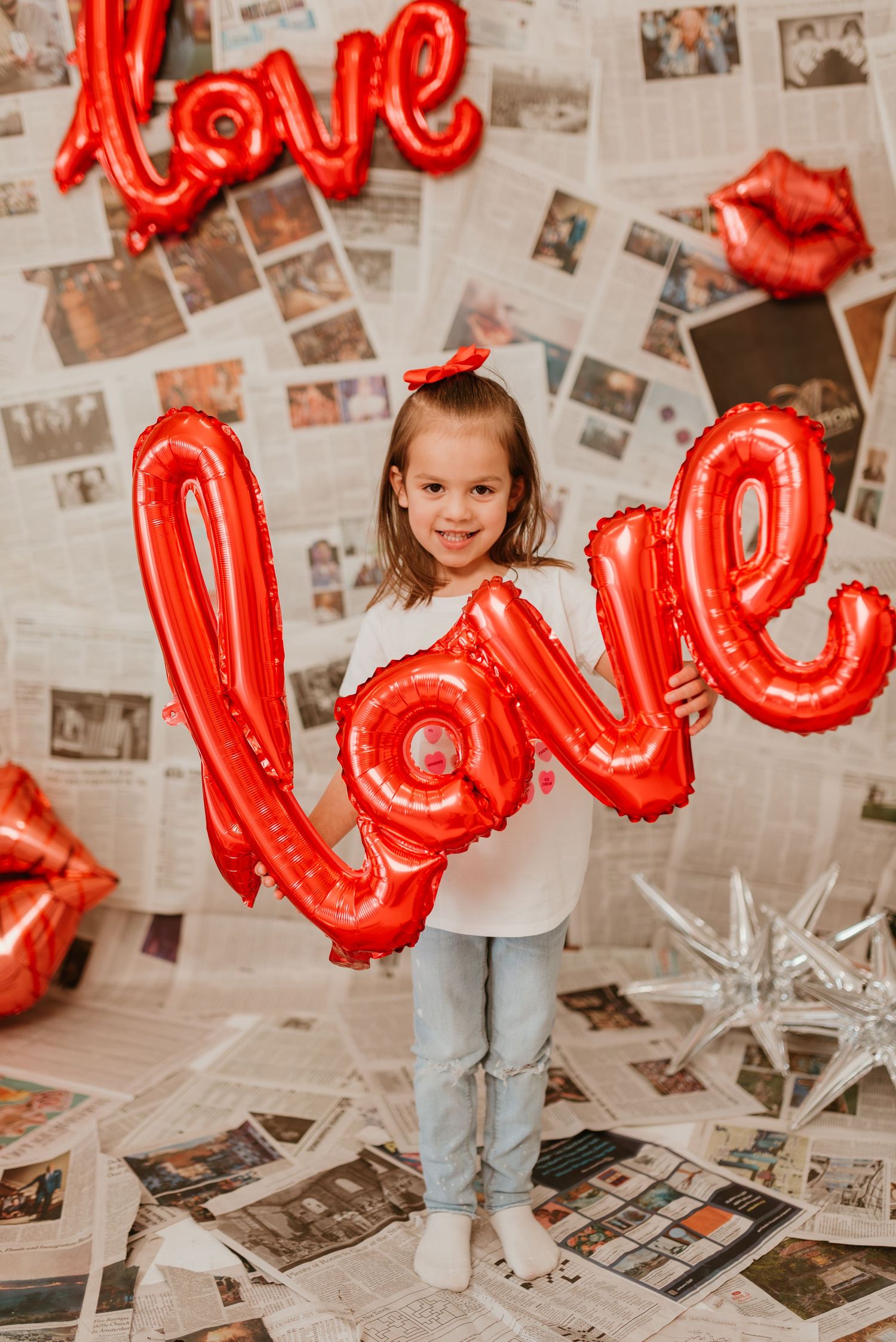 A smiling girl holds a red love balloon in front of a newspaper-covered wall with festive decor.