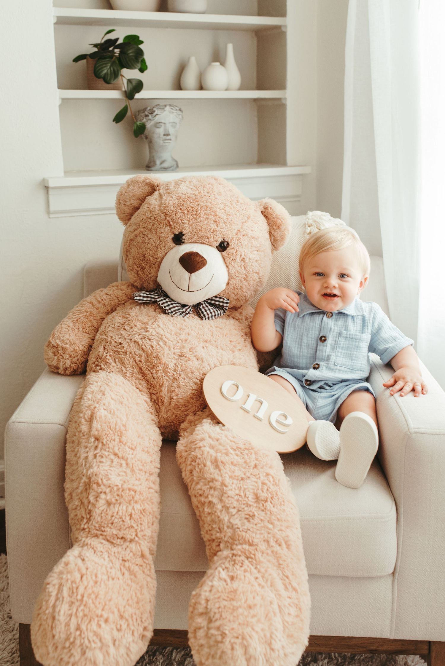 Smiling baby boy in blue romper sitting beside giant teddy bear holding wooden \
