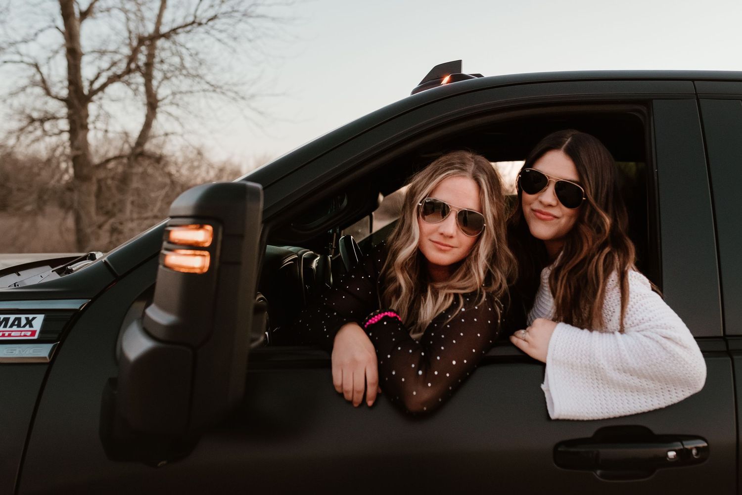 Two women wearing sunglasses lean out of a black truck window smiling during a golden hour photo shoot.