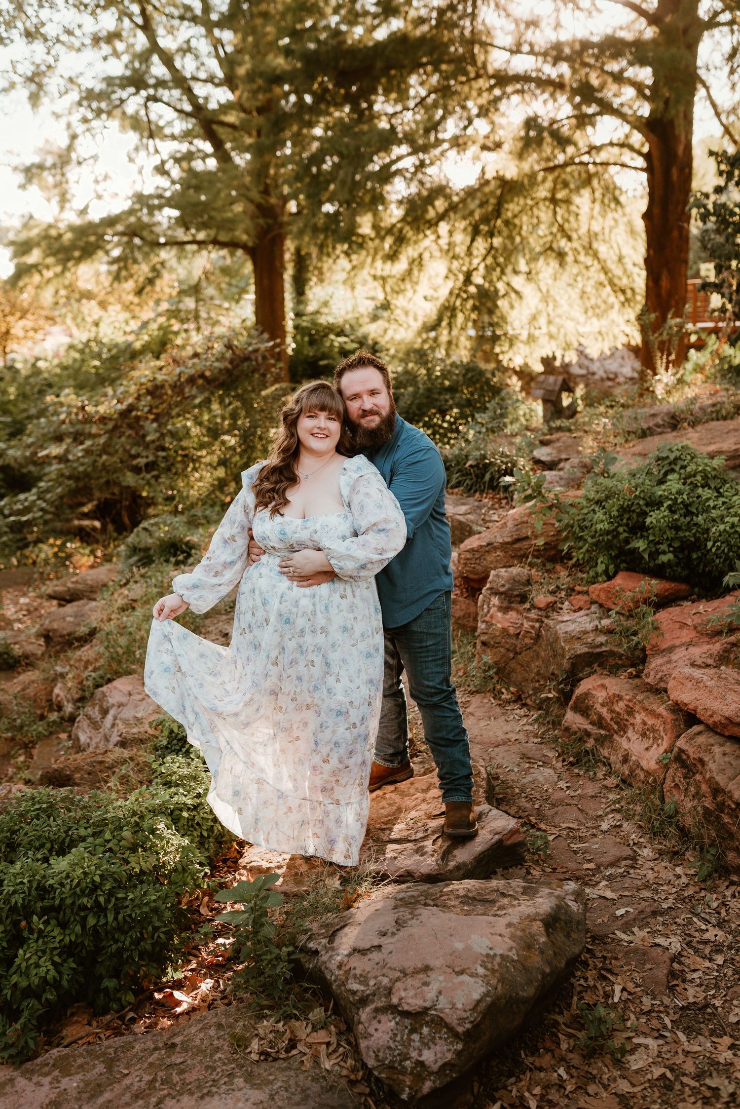 A couple poses on rocky terrain surrounded by trees, woman in a flowing floral dress.