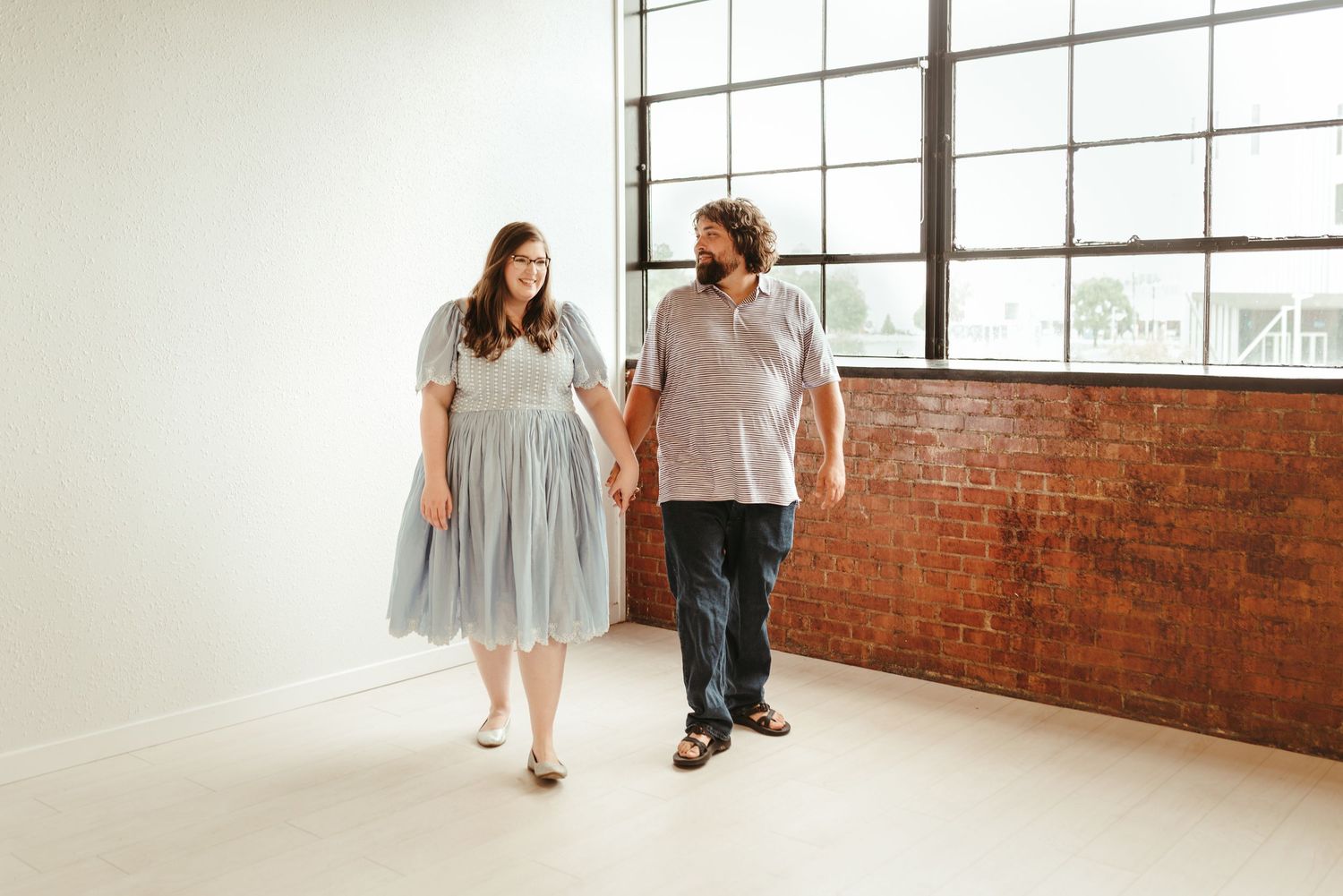 Couple holding hands and walking together in bright open space near brick wall and windows.