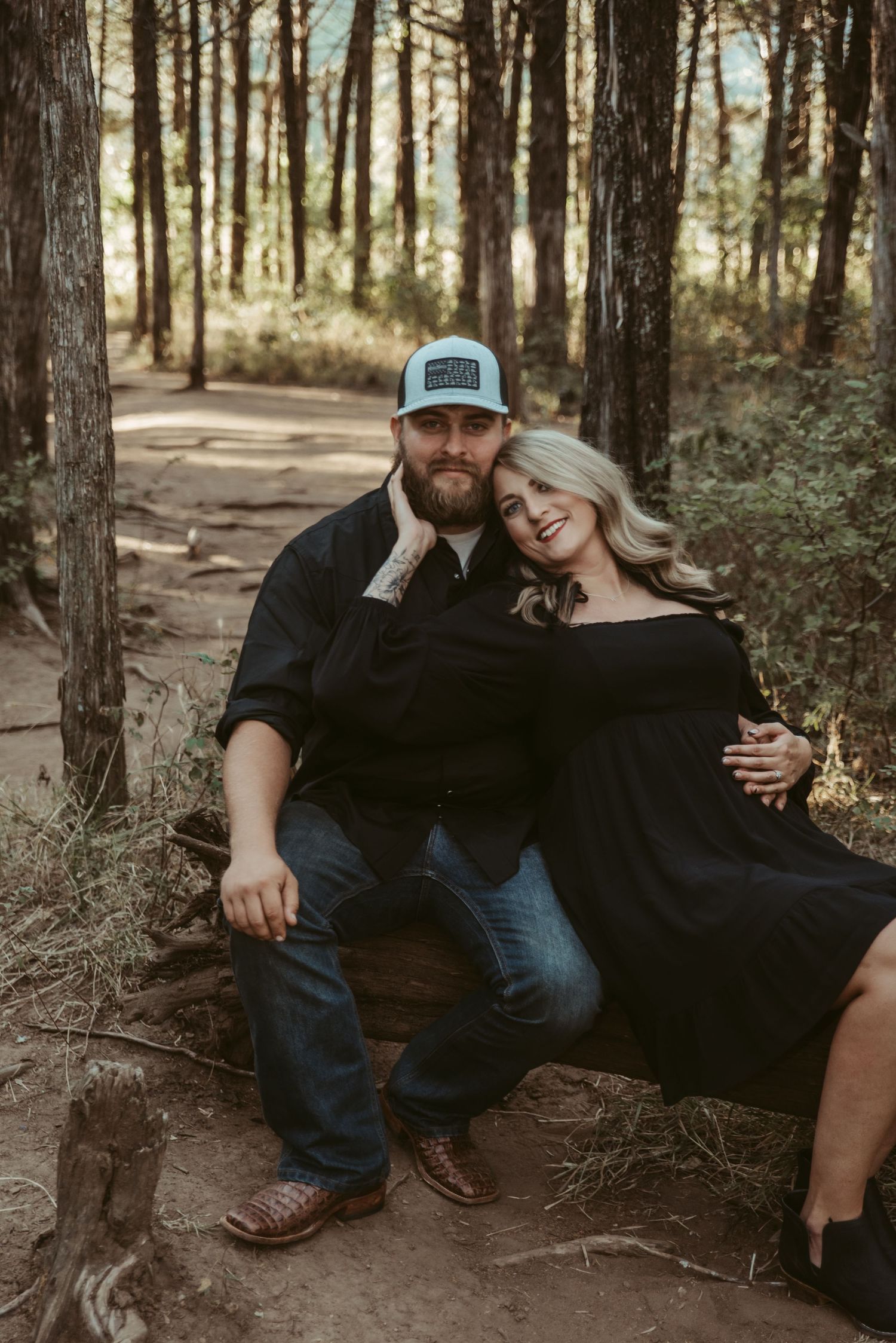 Couple sitting on a log in a pine forest, man in trucker hat and woman in black off-shoulder dress.