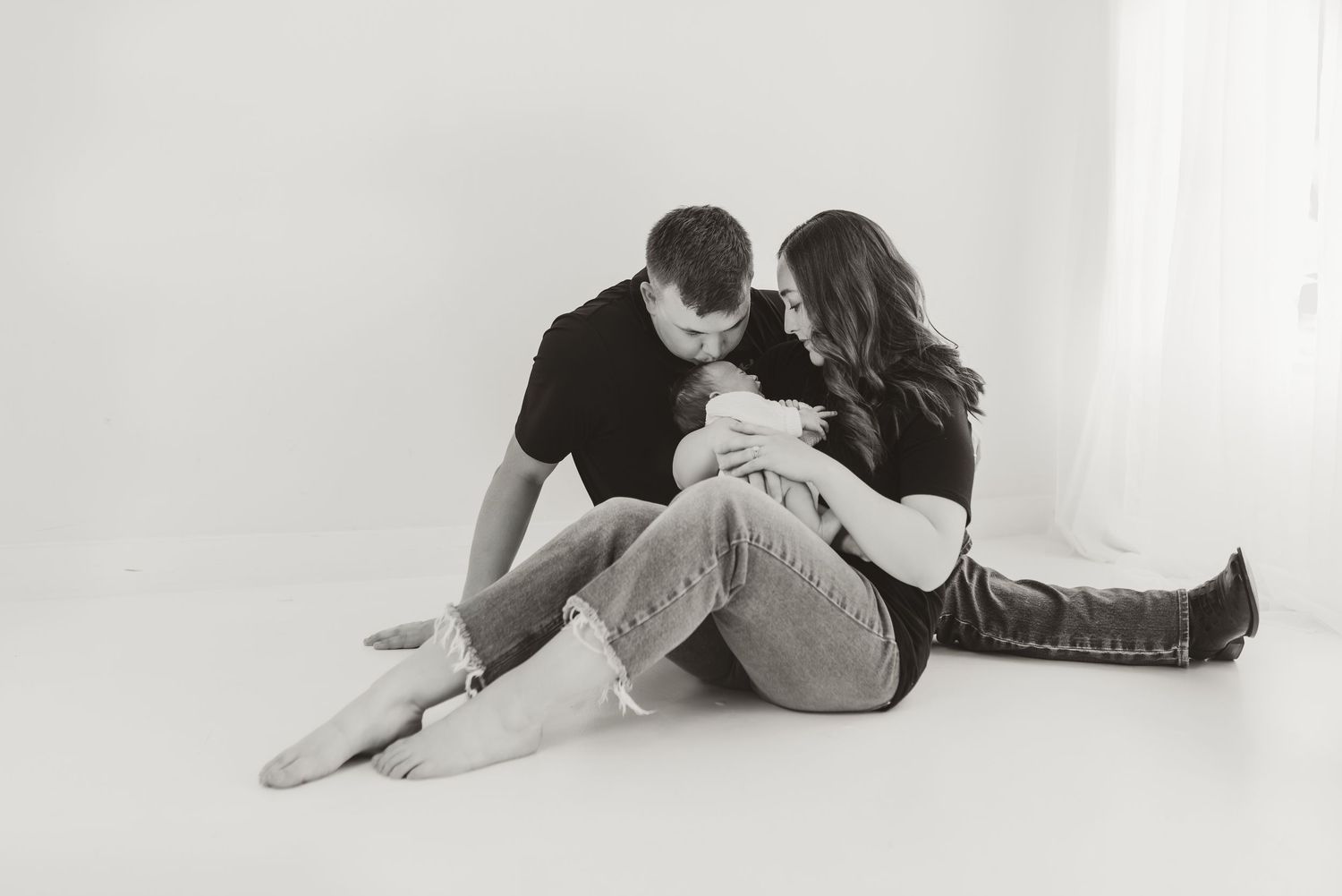 Black and white studio portrait of couple sitting on floor tenderly holding newborn baby together.