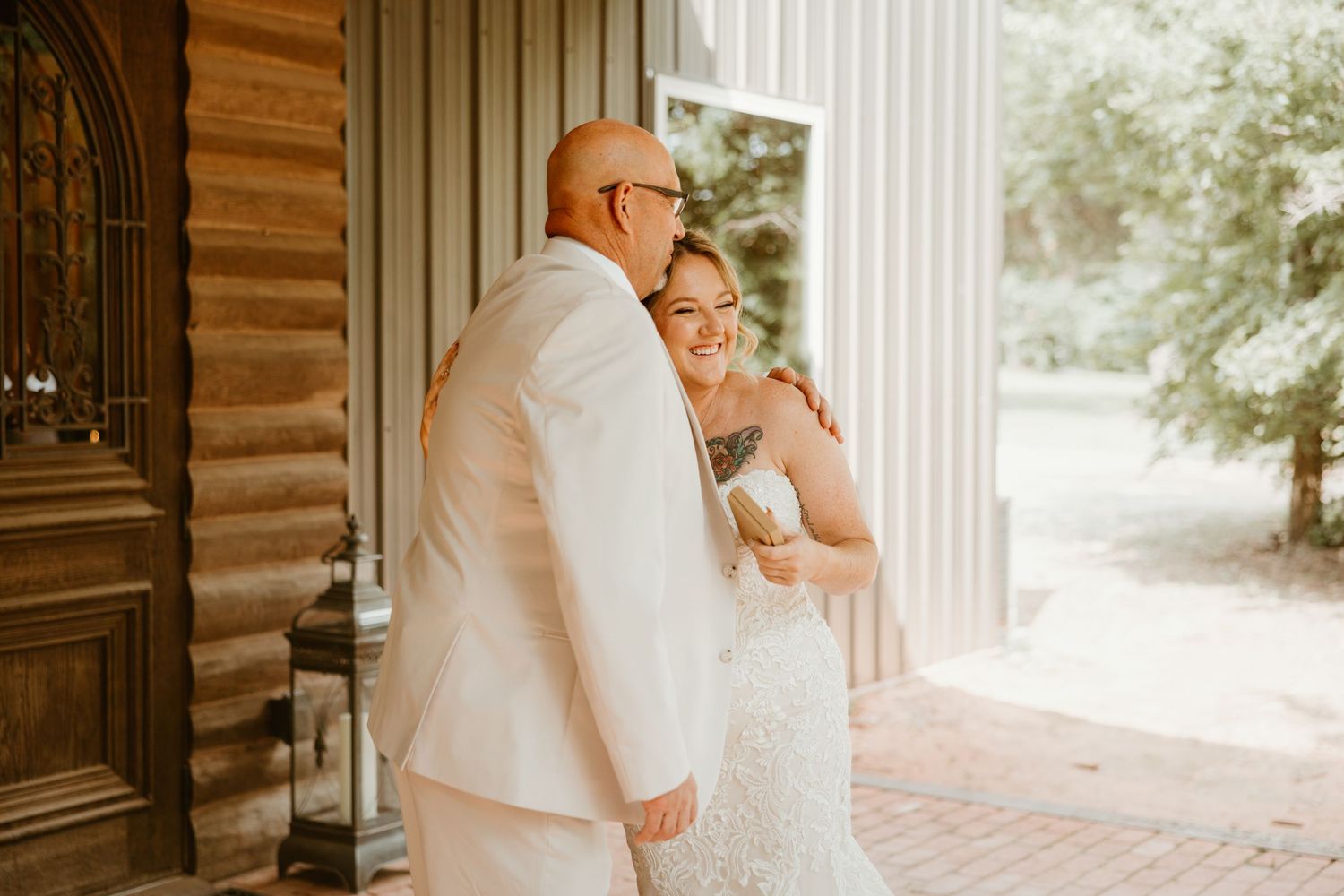 Bride and bald groom sharing a joyful embrace outside a rustic wooden building on wedding day.