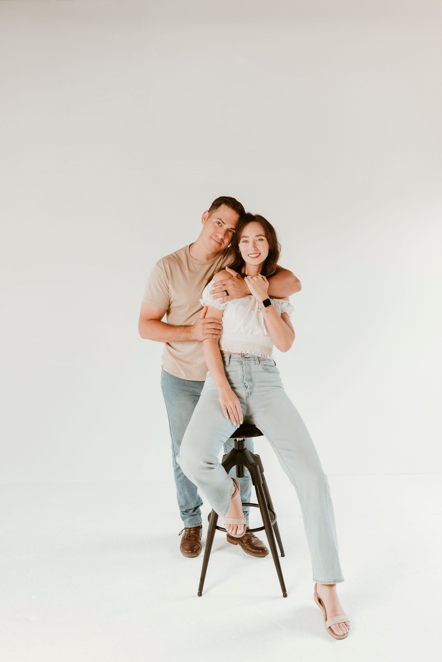 A smiling couple poses together on a stool against a clean white studio background.