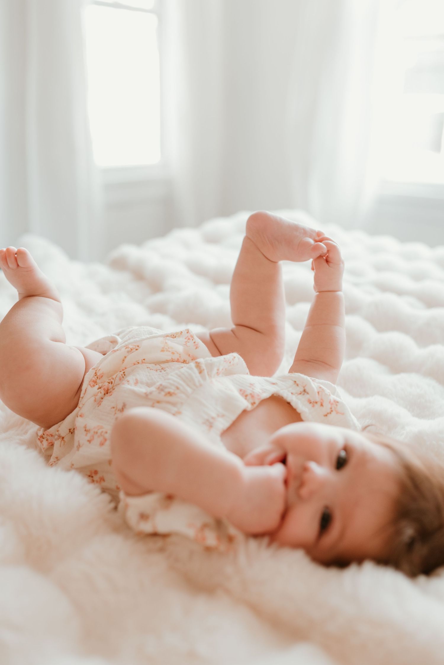 Adorable baby girl in floral romper laying on white fluffy blanket smiling and playing with her feet.