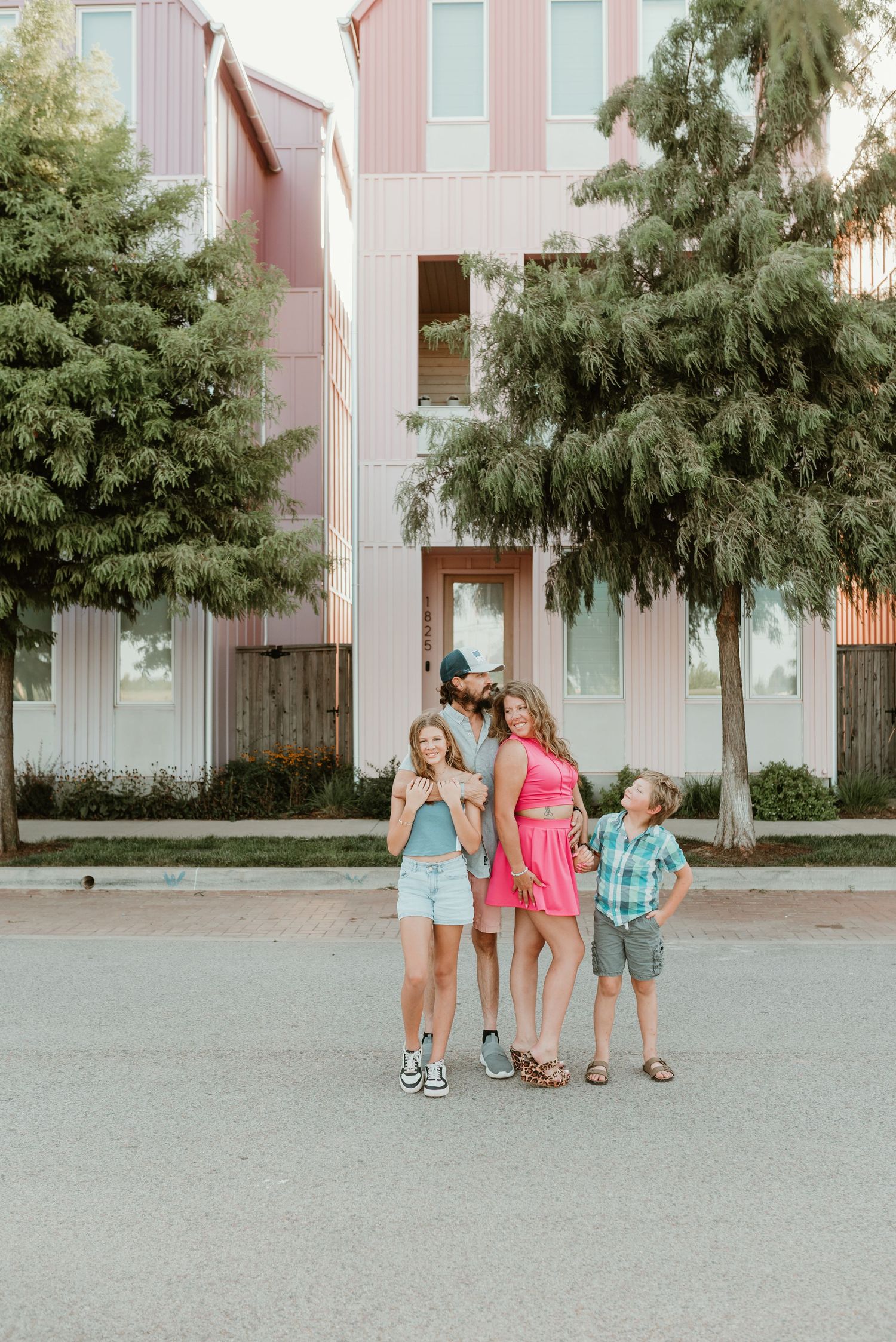 Family of four laughing together on a street in front of a modern pink building.