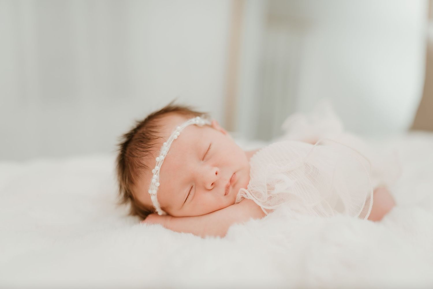Sleeping newborn baby girl in pink outfit with delicate white floral headband on white fur.