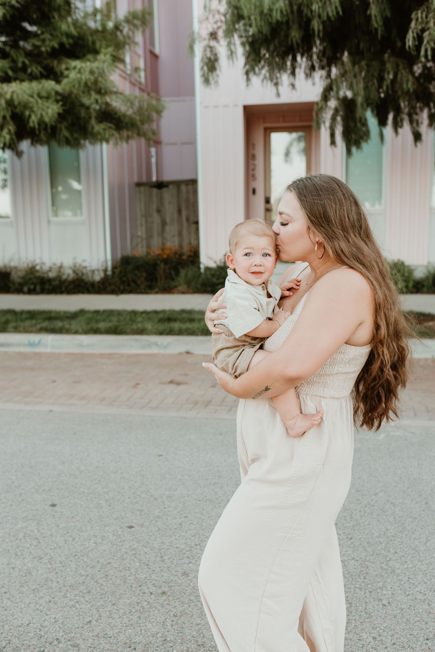 A young mother in a cream dress kisses her baby boy while standing outdoors near a modern building.