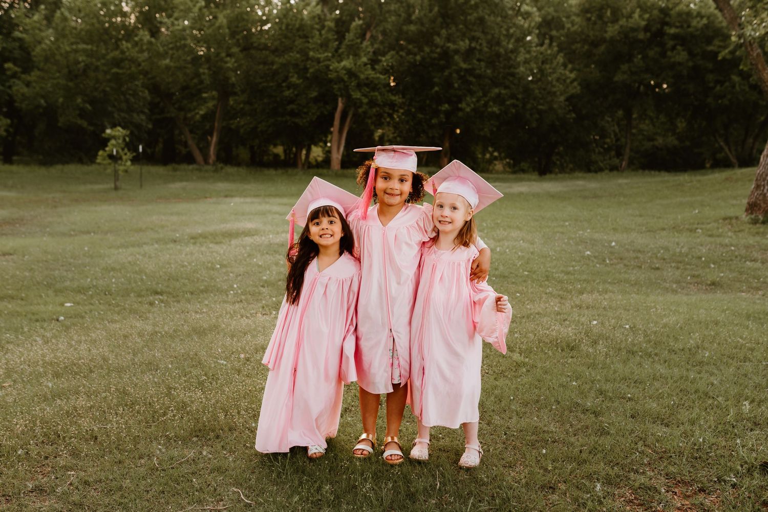 Three young girls in matching pink graduation caps and gowns standing together on green grass.