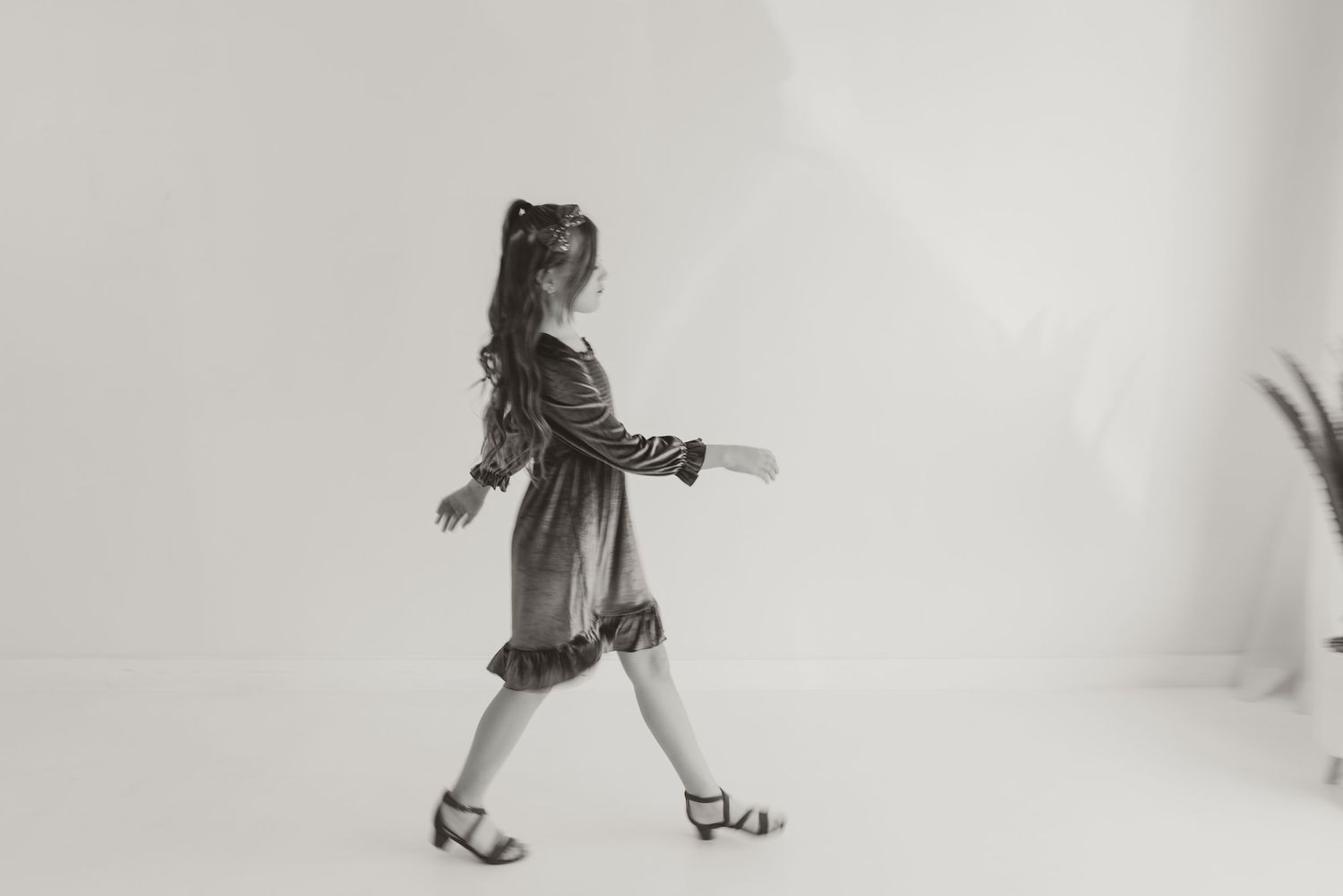 A young girl in a striped dress and heels walks gracefully in a bright white studio in black and white photo.