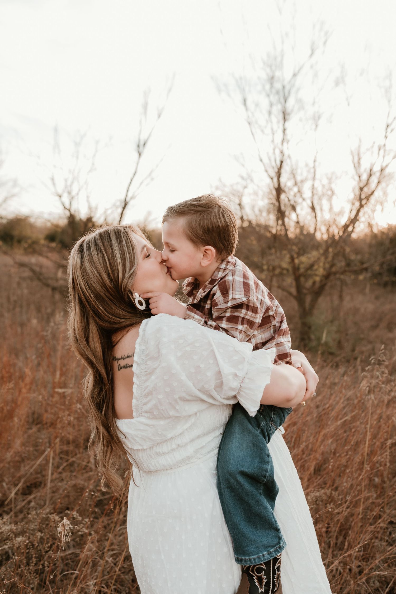 Pregnant woman in white lace dress kisses young son she holds in outdoor field setting.