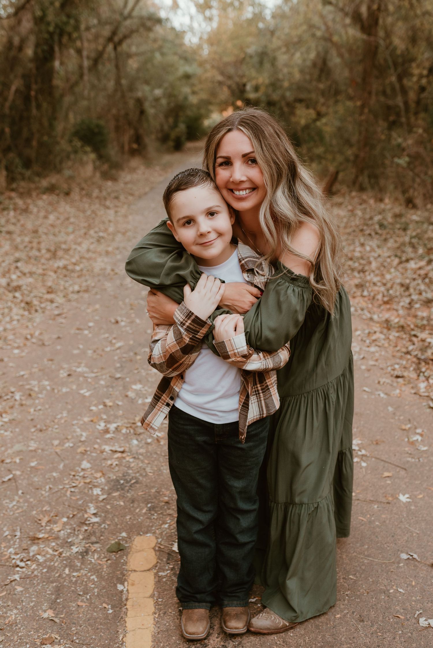 Young boy hugs smiling woman in green dress on a leaf-covered outdoor pathway.