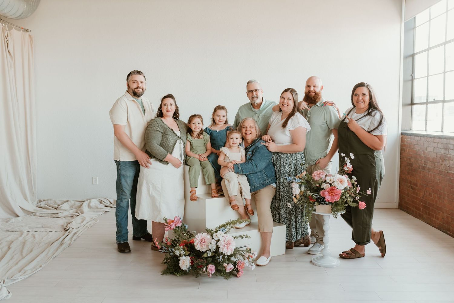 Extended family of nine adults and children pose together in bright studio with floral decor.