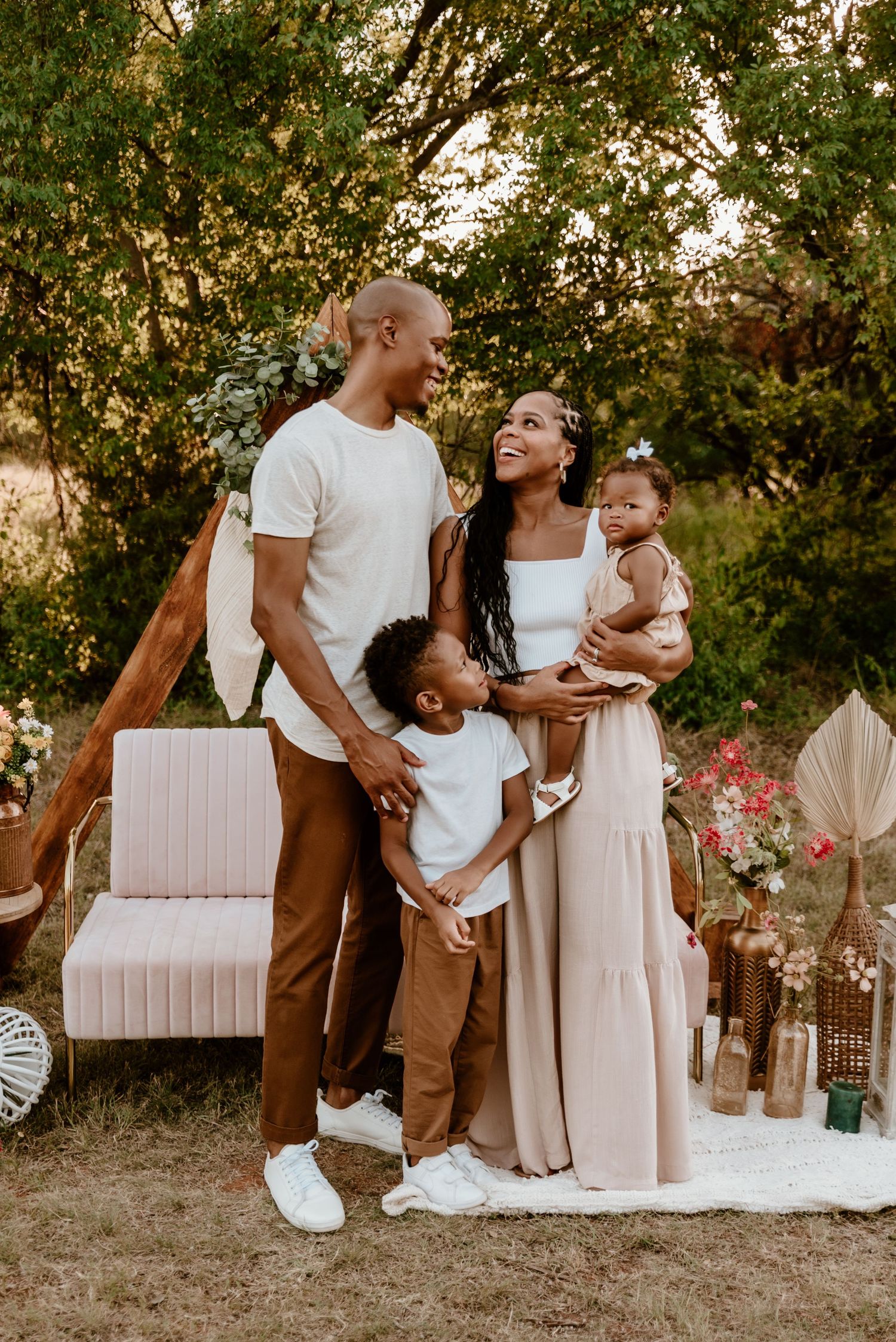 Black family of four poses outdoors at boho-styled photo session with triangular arch and floral decor.