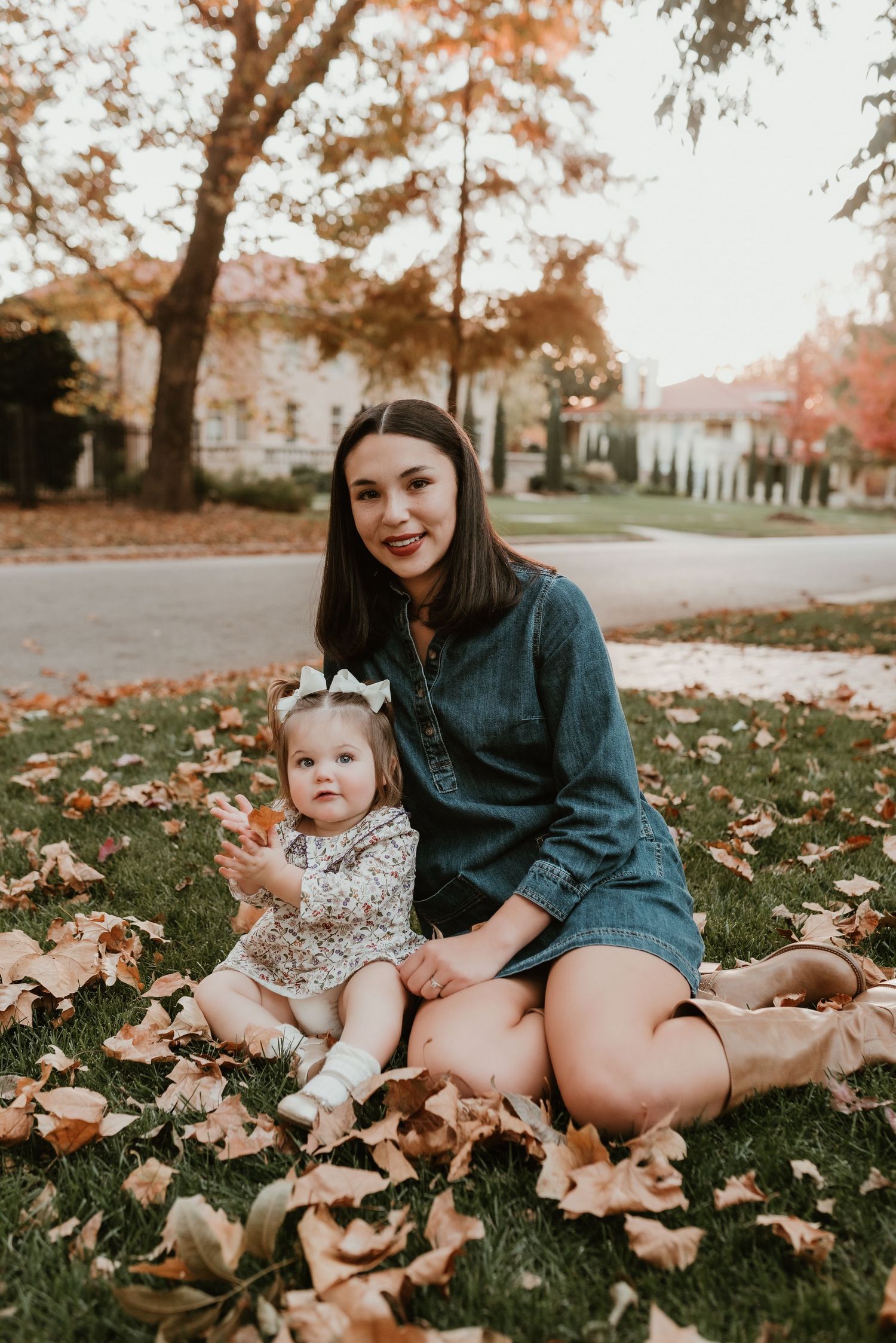 Young mother in denim dress sits on autumn grass with baby girl surrounded by fallen orange leaves.