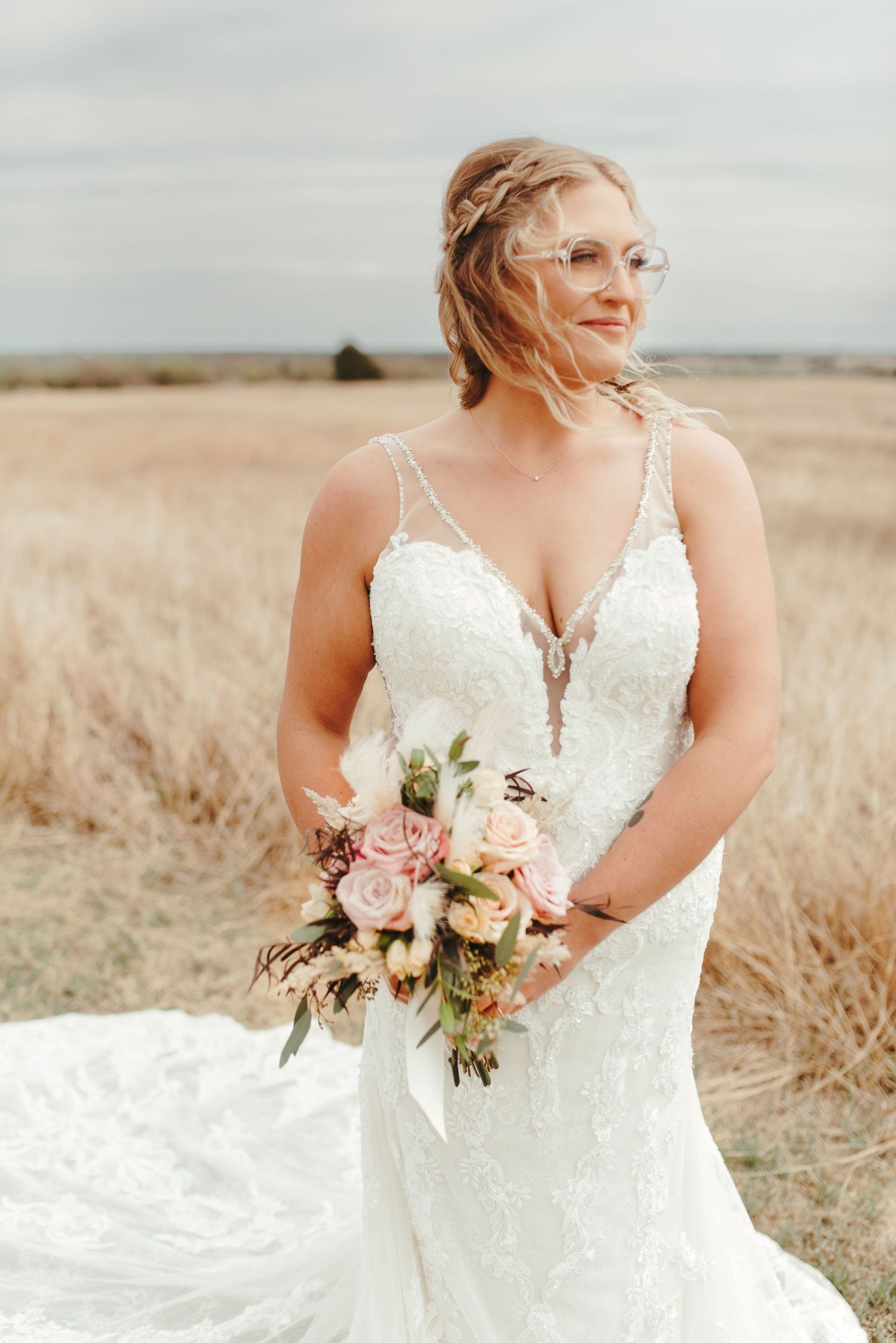 Bride in lace mermaid gown holds pink rose bouquet while standing in a golden field on her wedding day.
