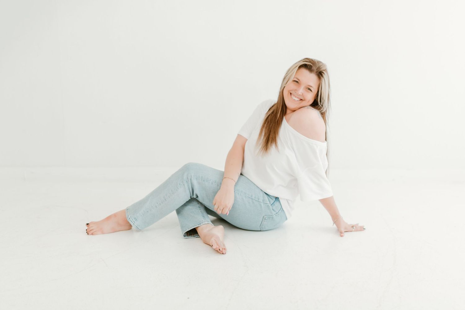 Smiling blonde woman sitting on white studio floor in light jeans and white off-shoulder top, barefoot.