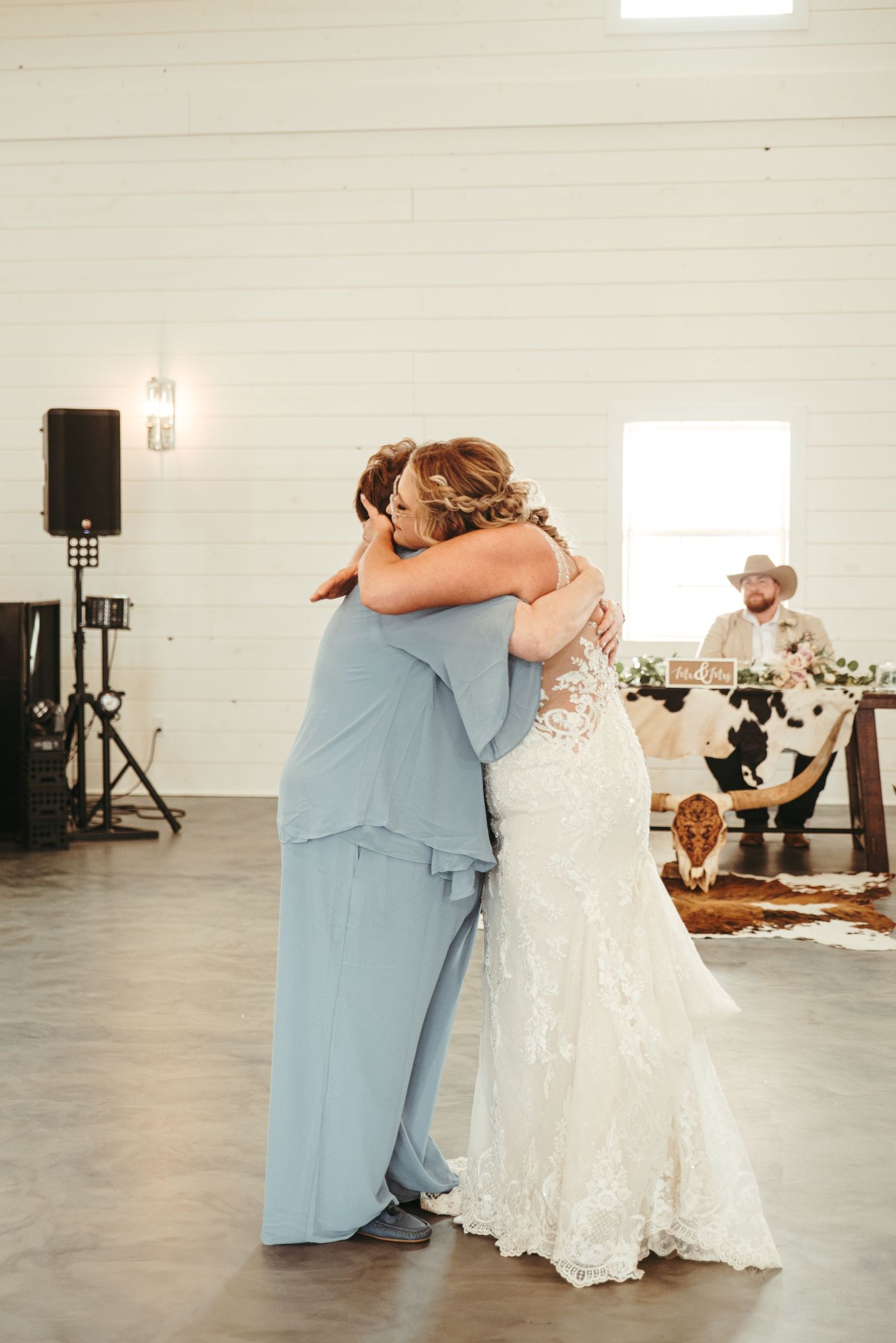 Bride in lace gown shares emotional hug with woman in blue dress on dance floor at country wedding reception.