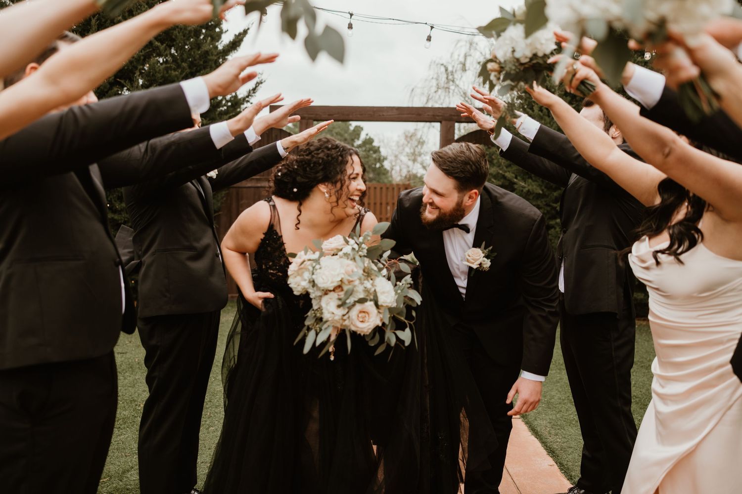 Wedding party creates an arch with their arms as bride in black gown and groom in tuxedo walk through outdoors.