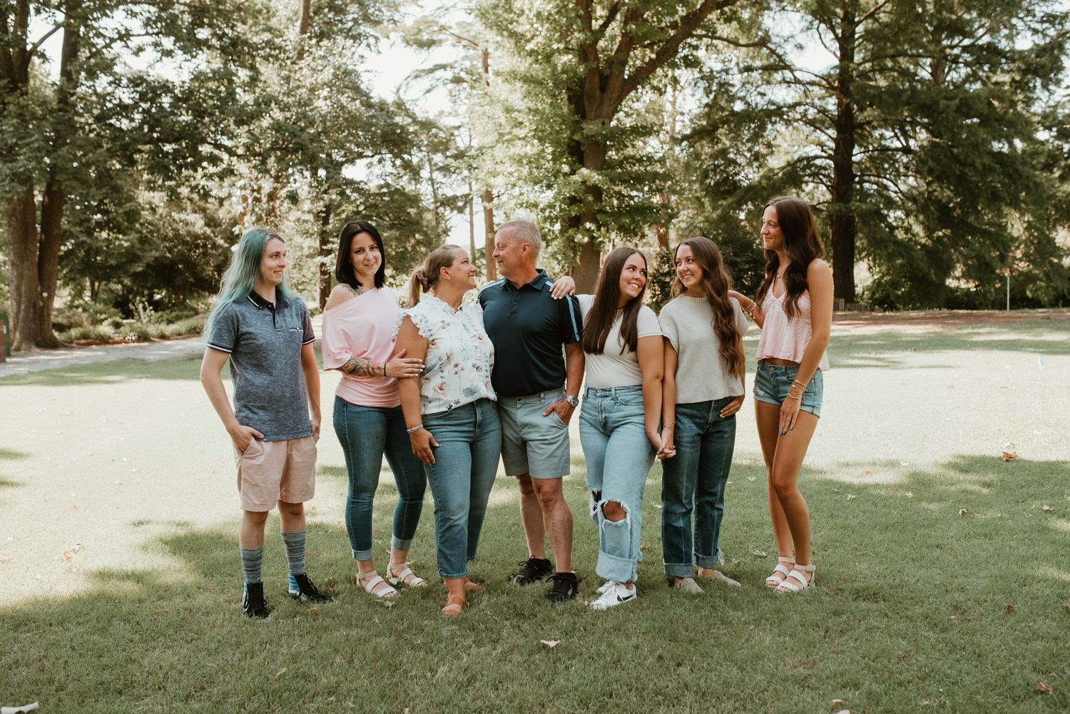 Family of seven smiling together outdoors in a tree-lined park on a sunny day.