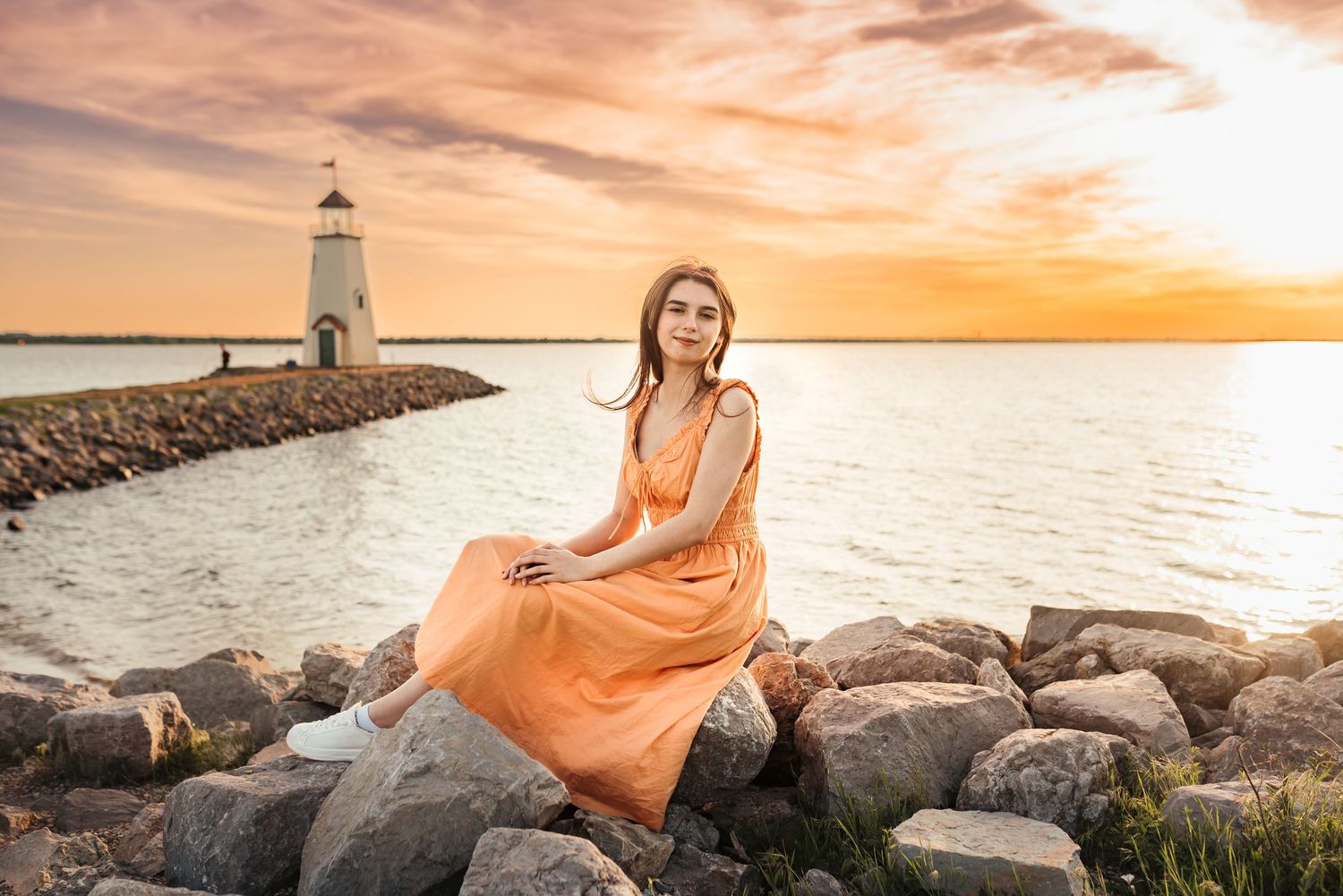 A young woman in an orange dress sits on rocks near a lighthouse at golden sunset.
