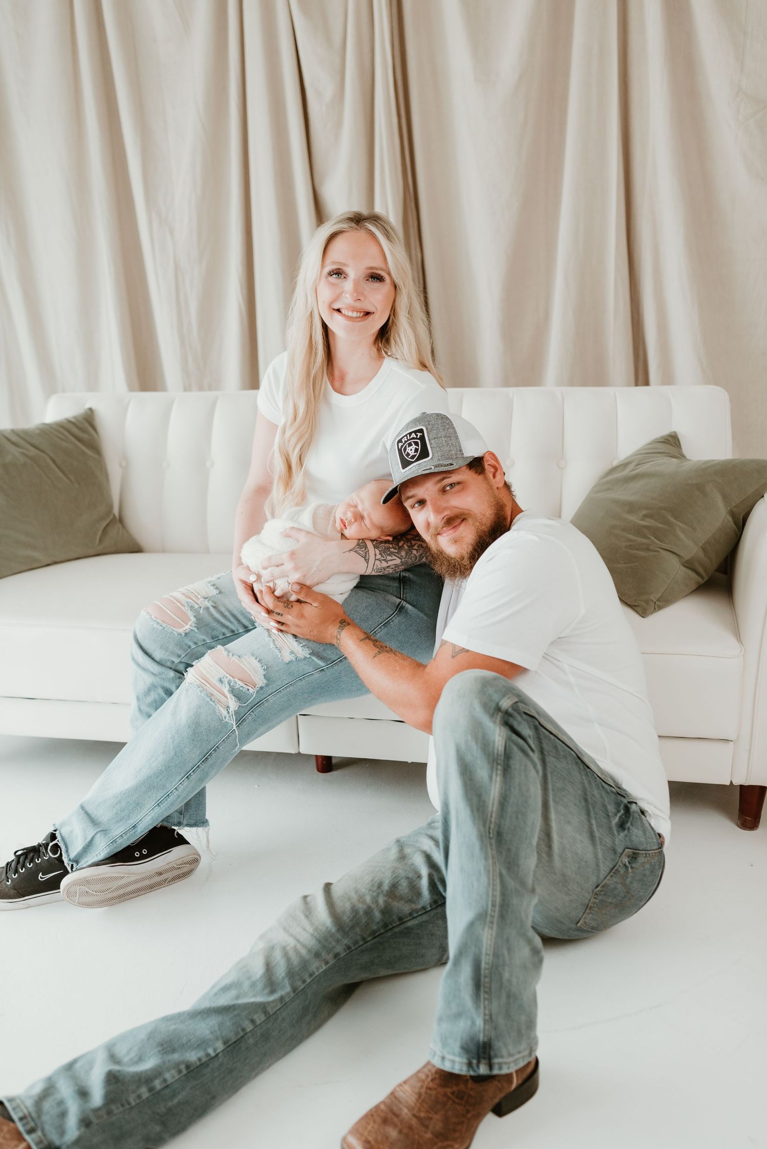 Smiling couple in white tees and jeans posing with their newborn baby on a white sofa.
