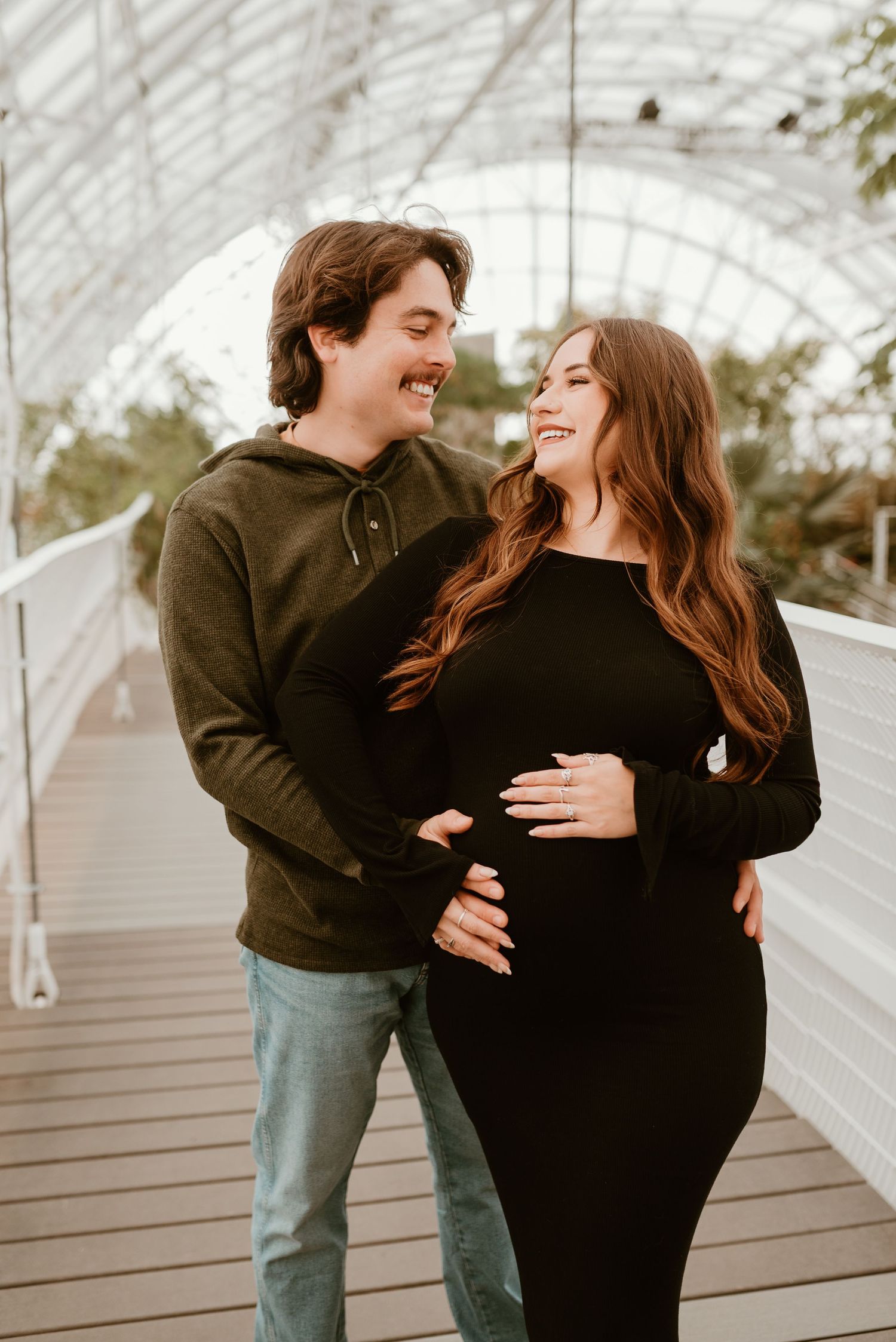 Happy couple laughing together in greenhouse, woman in black maternity dress holding baby bump.