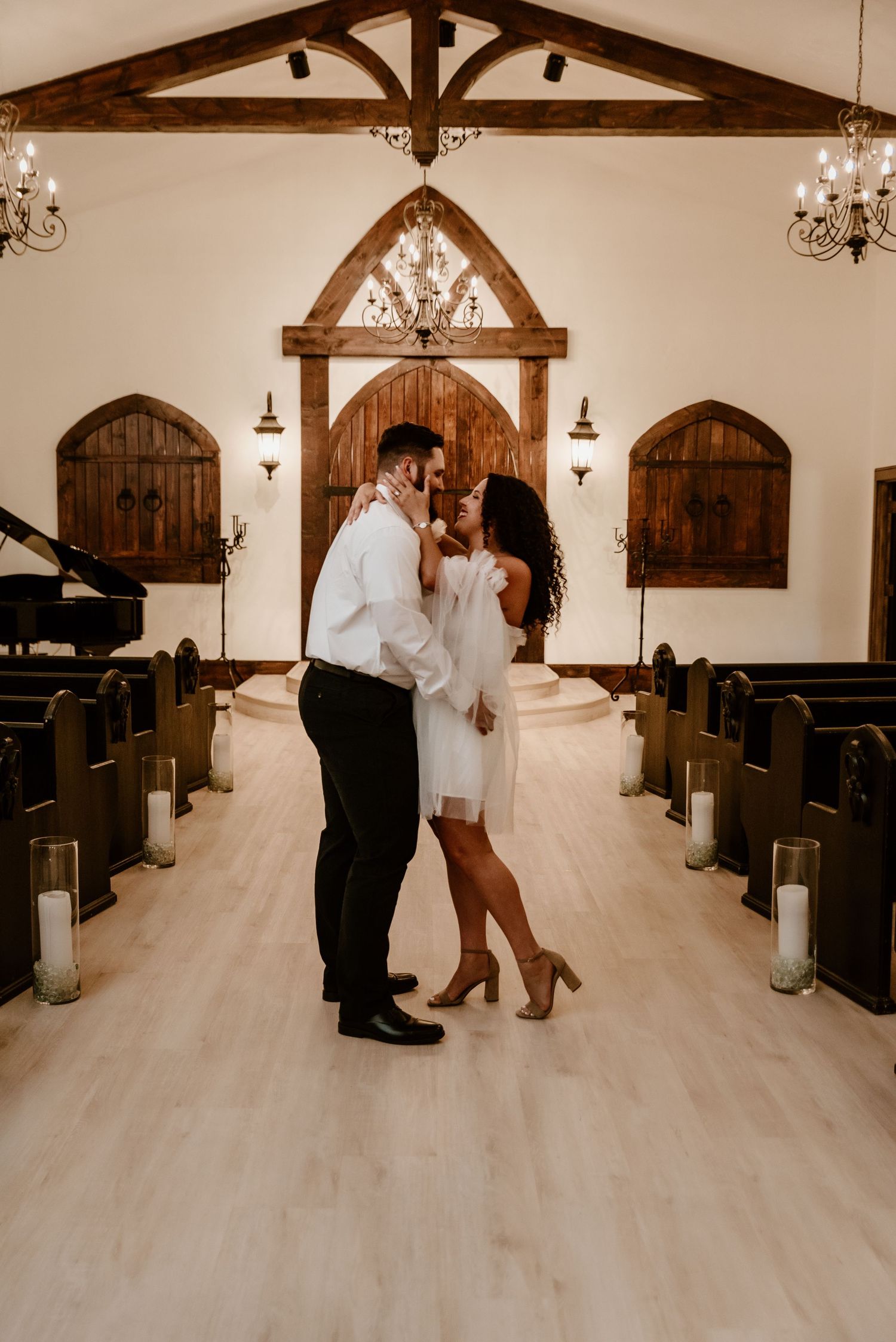 Couple sharing a romantic moment in an elegant white chapel with wooden arched details and chandelier lighting.