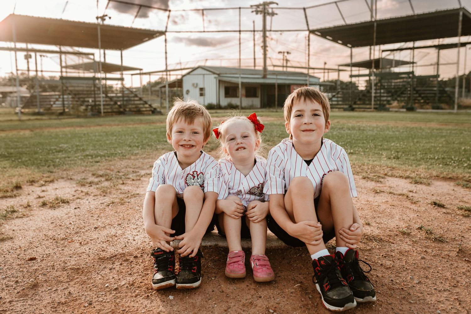 Three young siblings in baseball uniforms sitting together smiling on a dirt baseball field.