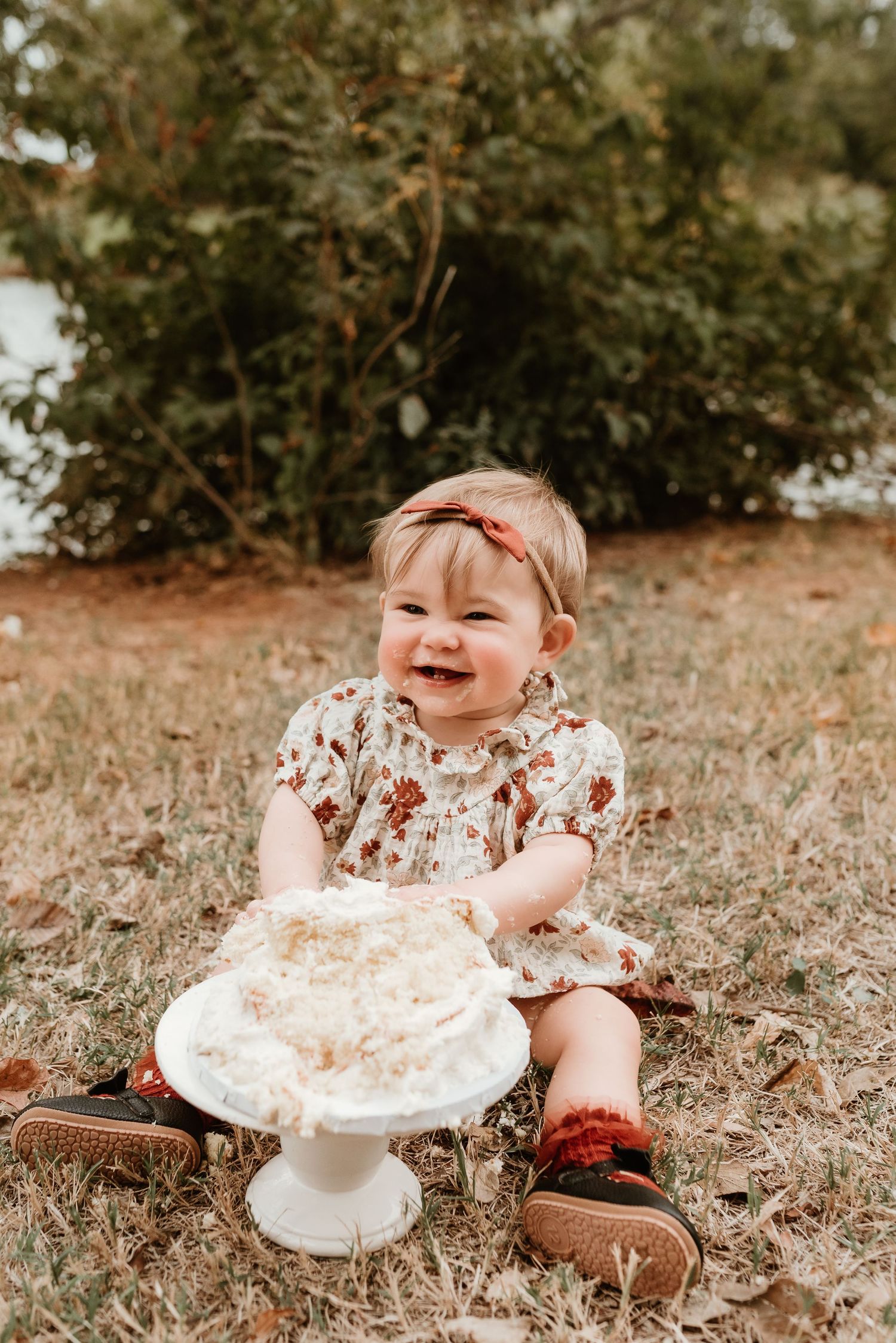Happy baby girl laughs joyfully while smashing her first birthday cake outdoors on grass.