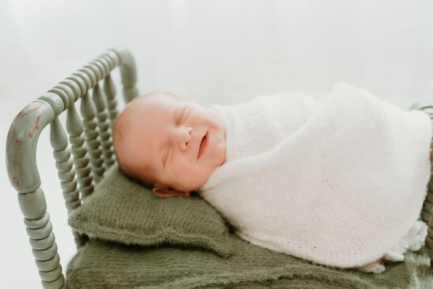 Smiling sleeping newborn baby wrapped in white swaddle lying in a small green wooden bed.
