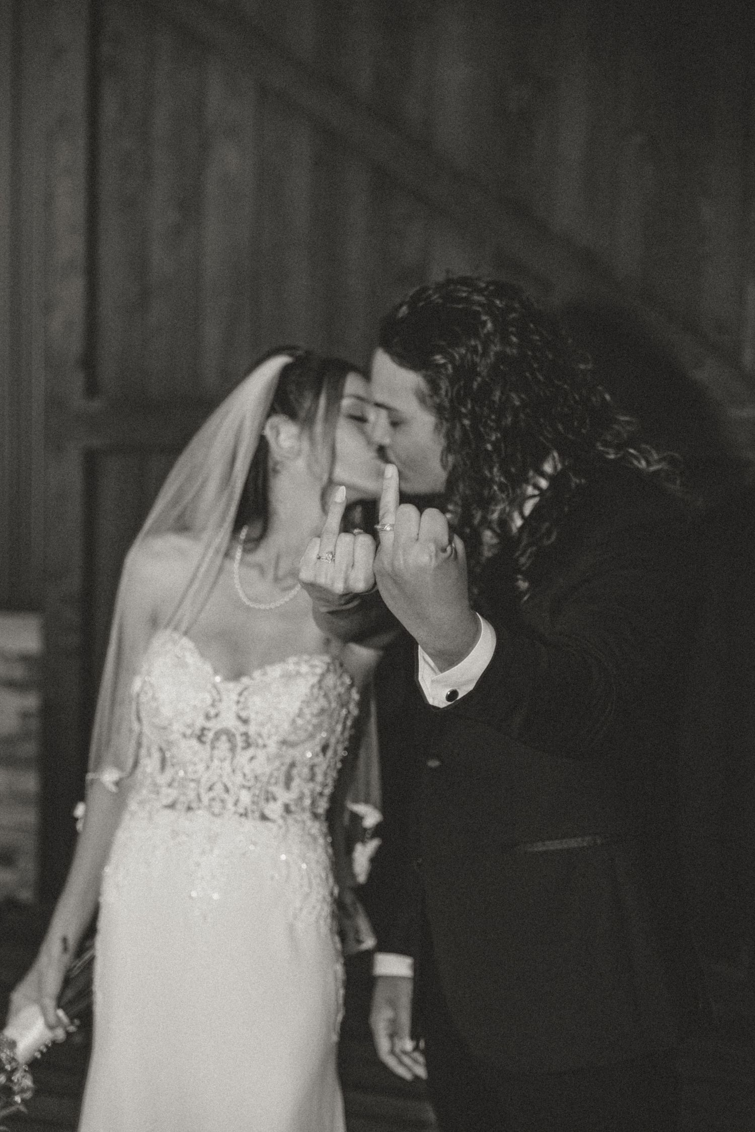Black and white photo of bride and groom sharing first kiss while displaying wedding rings at barn wedding venue.