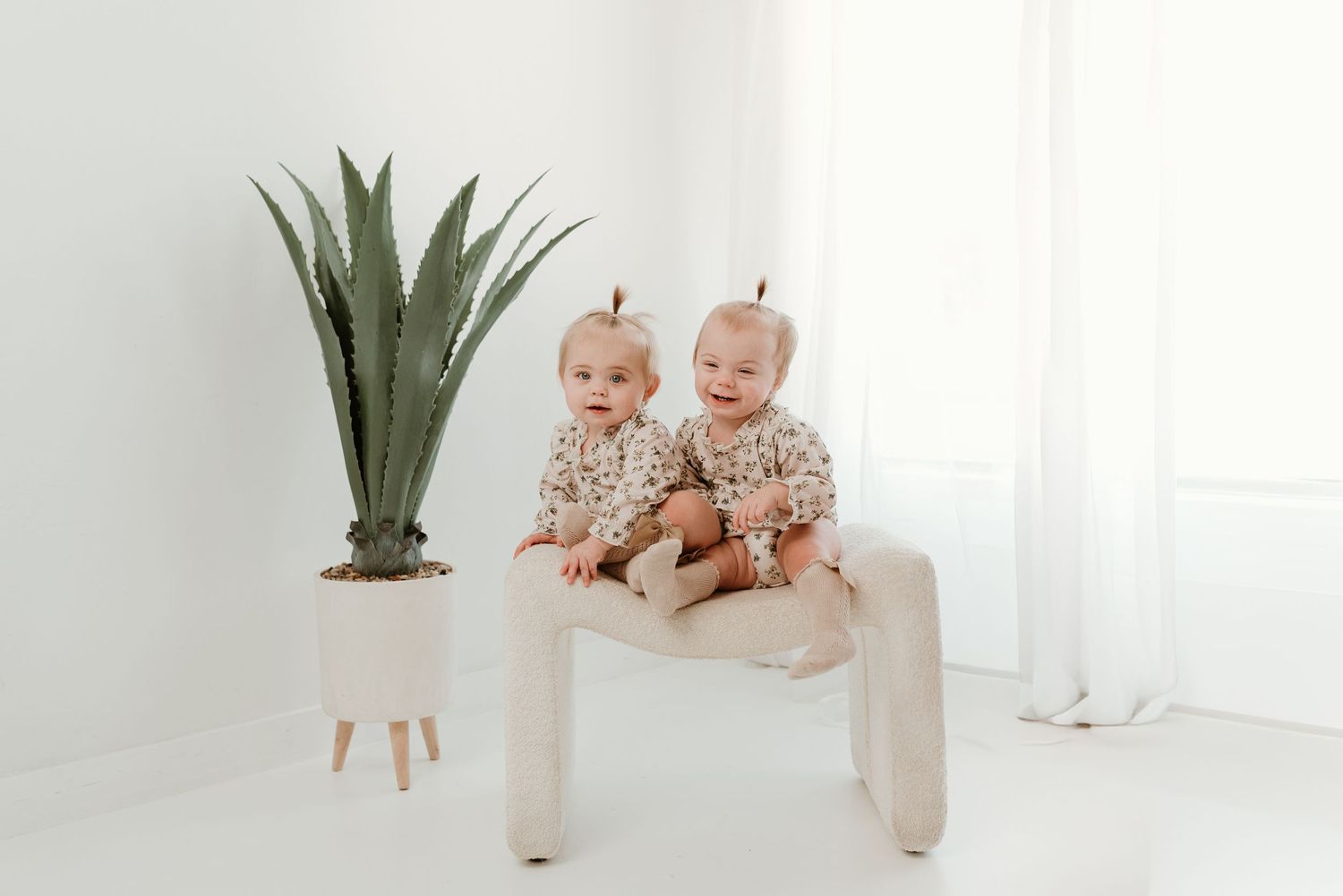 Adorable twin babies in floral outfits sitting together on a white bench beside a potted plant.