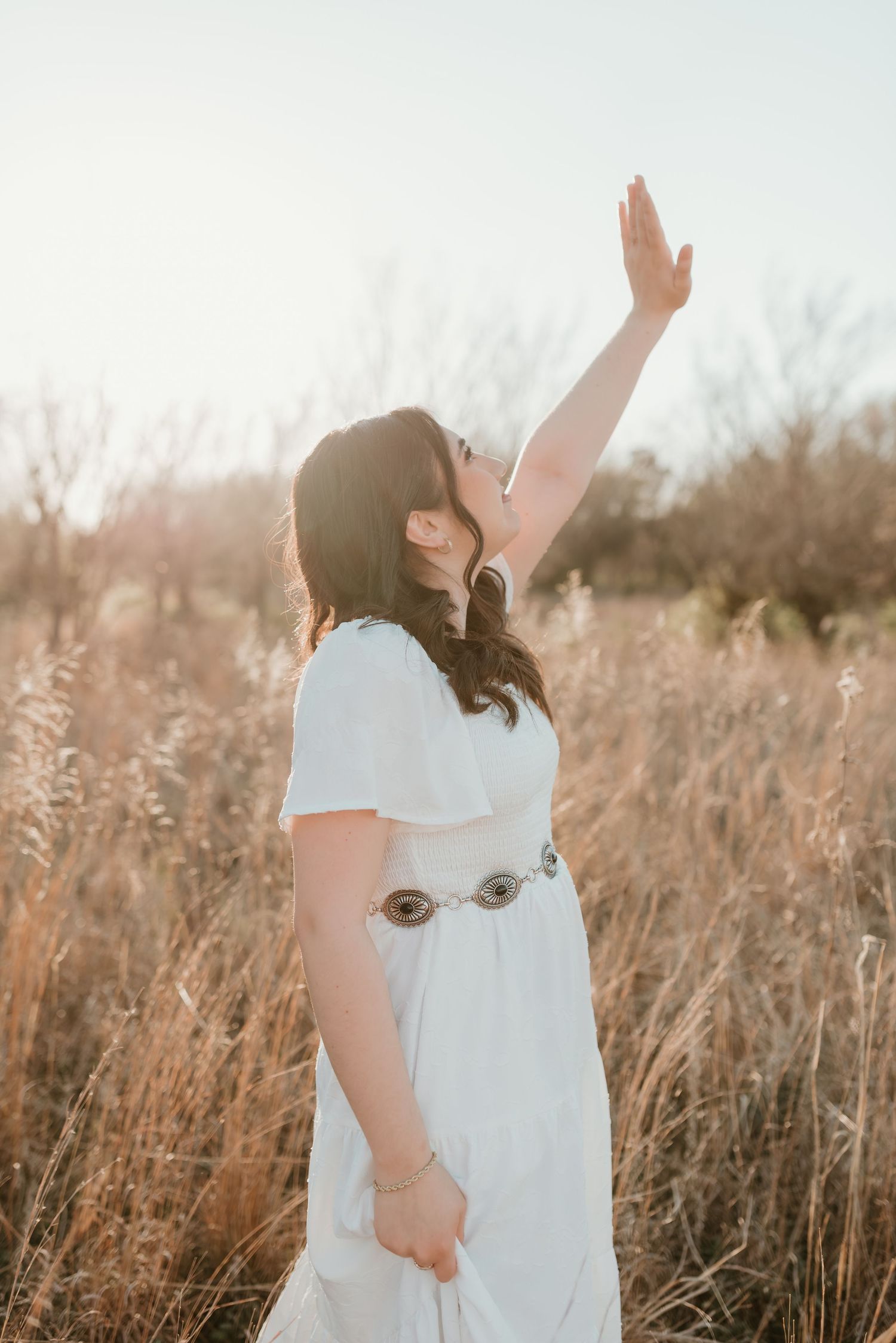 Woman in white dress with concho belt raising hand toward sky in golden hour sunlit field.