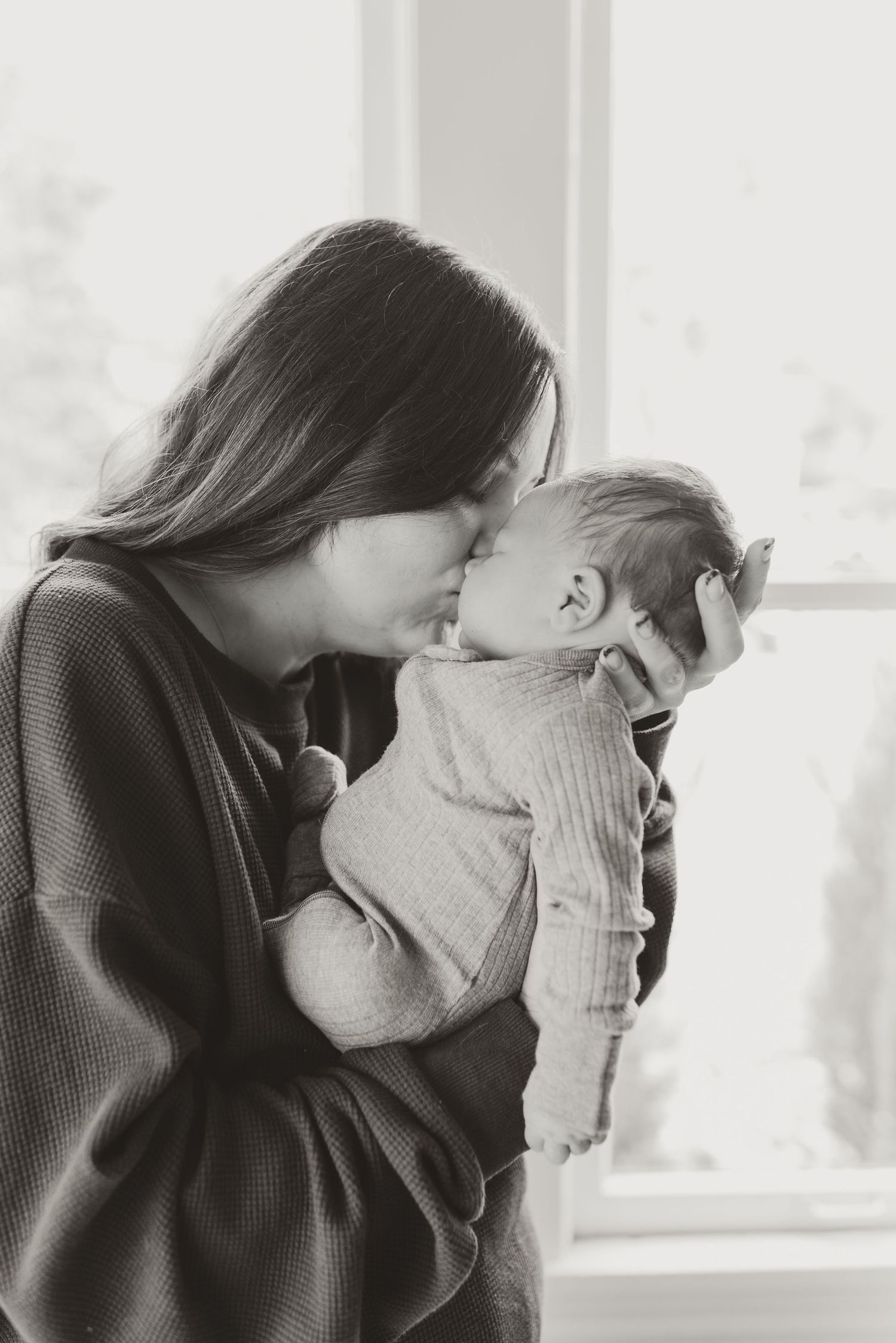 Tender black and white photo of a mother gently kissing her newborn baby by a bright window.