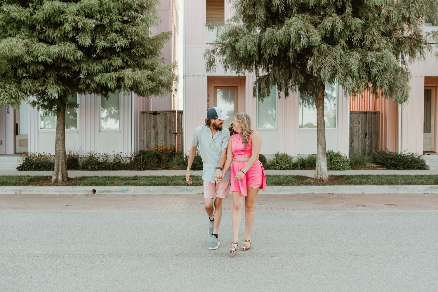Happy couple walking hand in hand on a sunny street with trees and modern building behind them.
