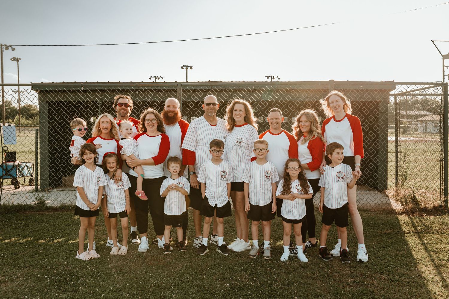 Family baseball team photo in red and white uniforms on outdoor baseball field at sunset.