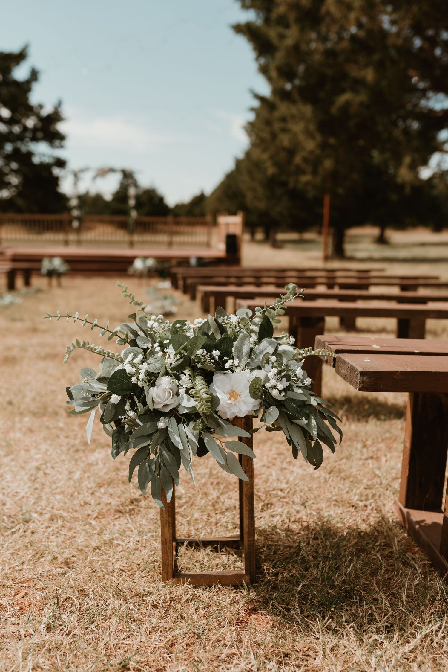 White floral arrangement with greenery on wooden stand decorating outdoor wedding ceremony aisle.