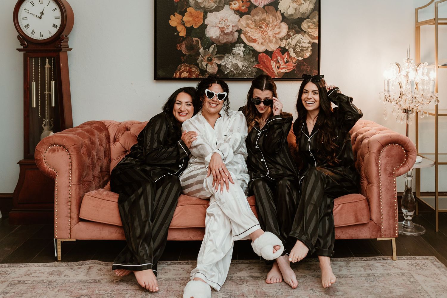 Four women in matching black satin pajamas pose together on a pink velvet sofa for a bridal party getting ready photo.