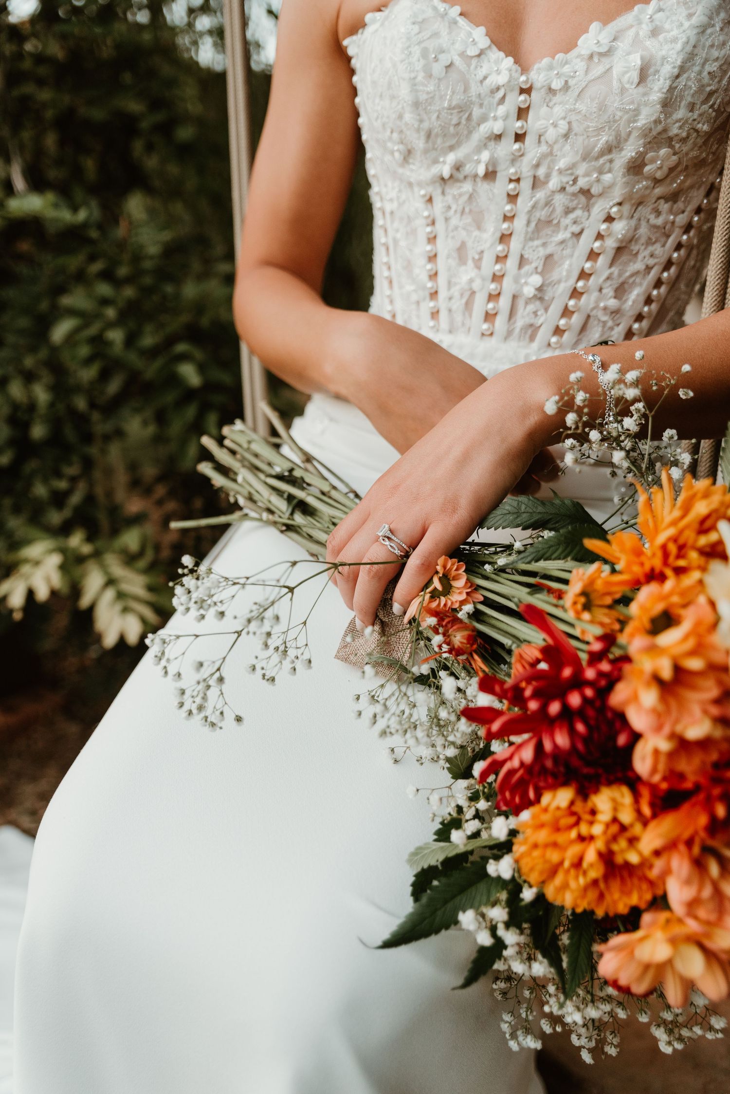 Bride in lace wedding dress holding vibrant orange and red floral bouquet with baby's breath outdoors.