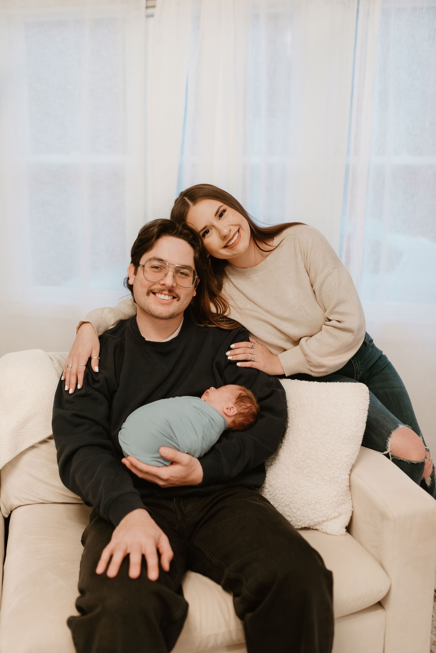 Smiling new parents sitting on white couch holding swaddled newborn baby in bright room.