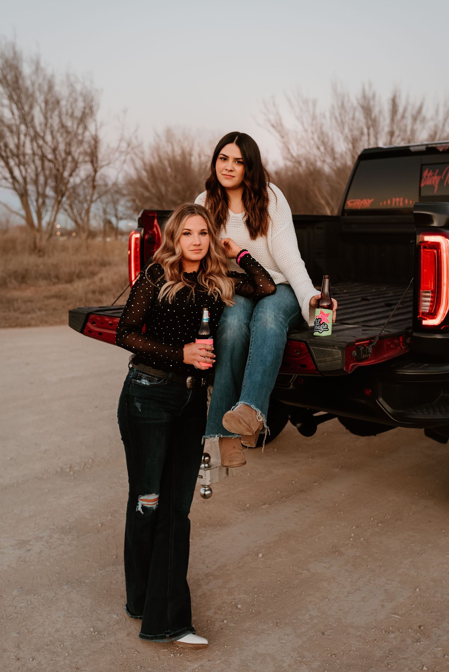 Two women pose by a black pickup truck tailgate holding beverages in a rustic outdoor setting at dusk.