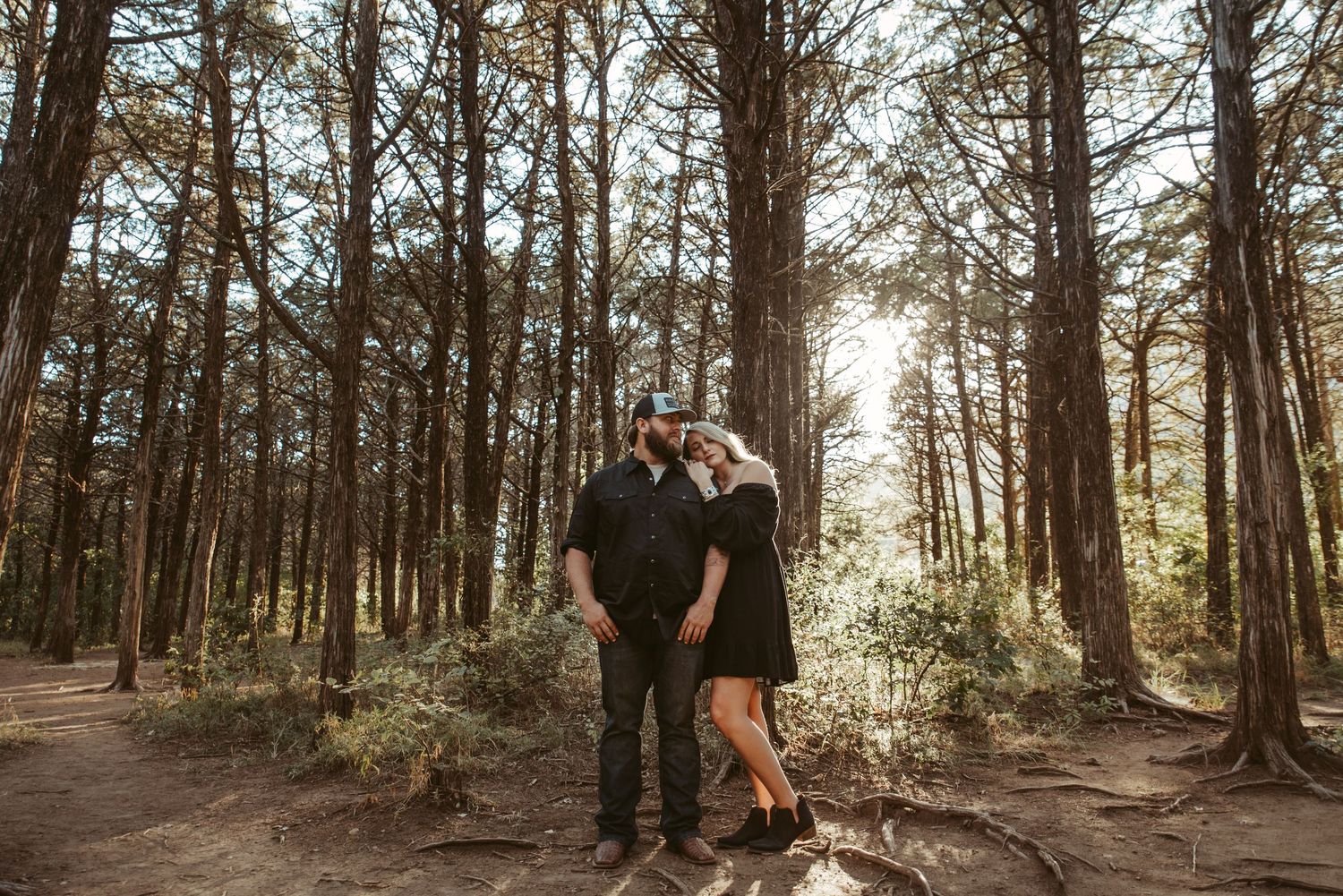 Couple in black outfits embracing in a sunlit pine forest during an outdoor engagement photo session.